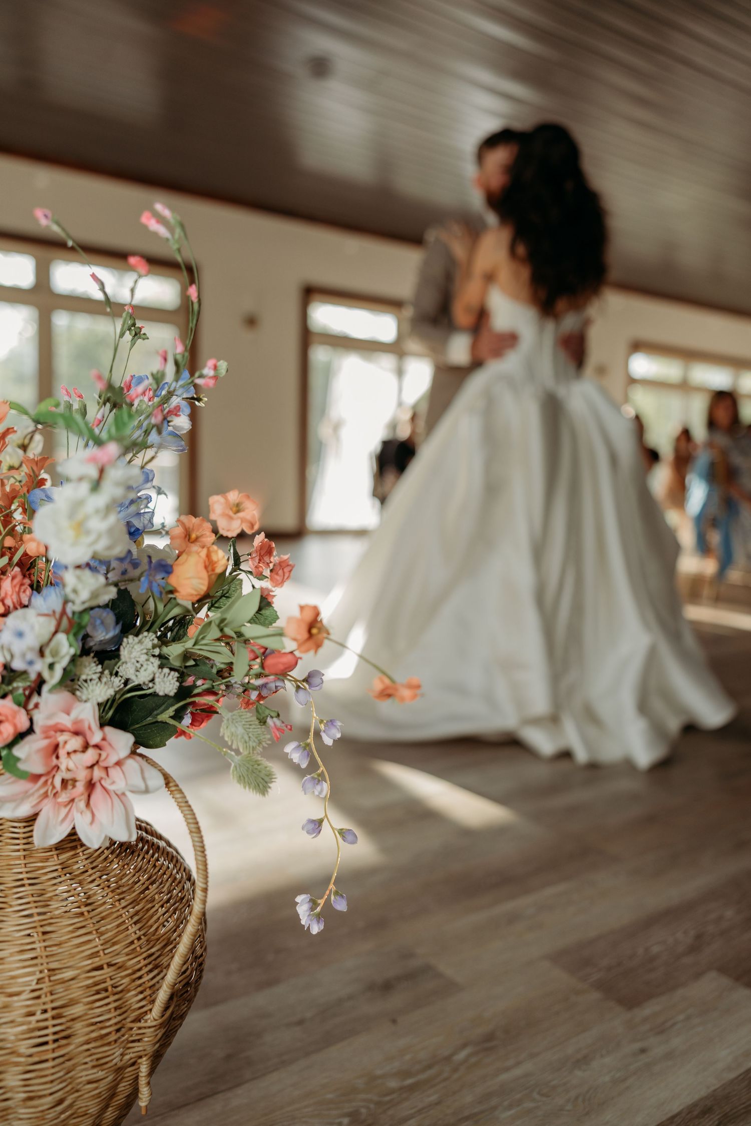 Colorful wildflower basket arrangement in foreground as bride and groom embrace during wedding reception.