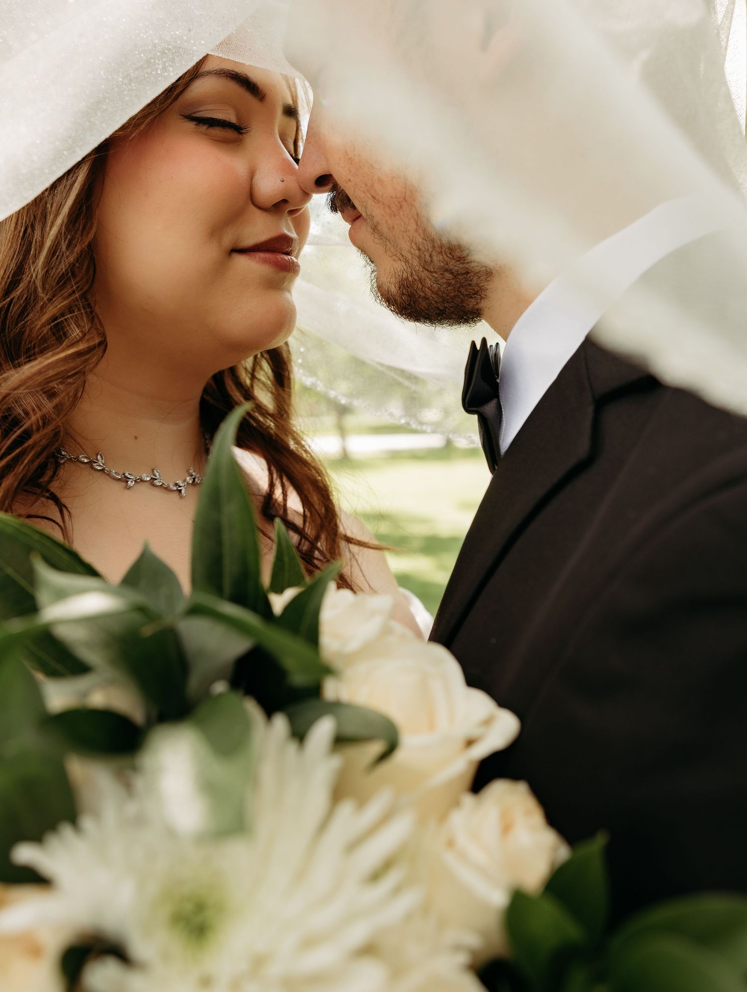 Bride and groom share an intimate nose-to-nose moment under a flowing veil, with a white floral bouquet in the foreground.