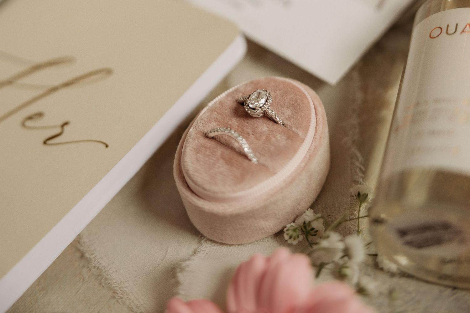 An elegant engagement ring sits in a pink velvet oval ring box beside delicate floral decor and paper.