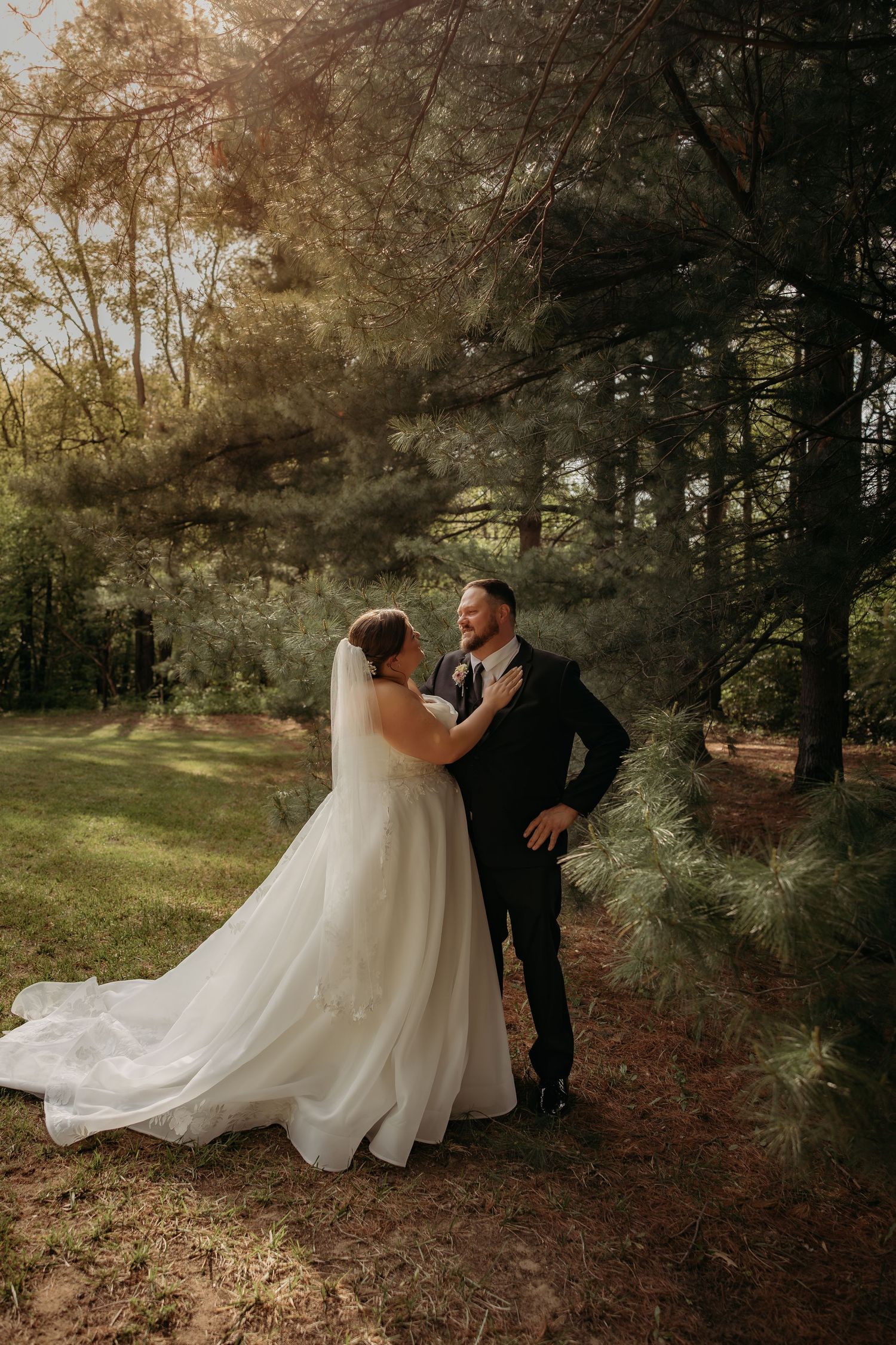 A bride and groom share a romantic moment among tall pine trees in golden sunlight during their outdoor wedding portrait.