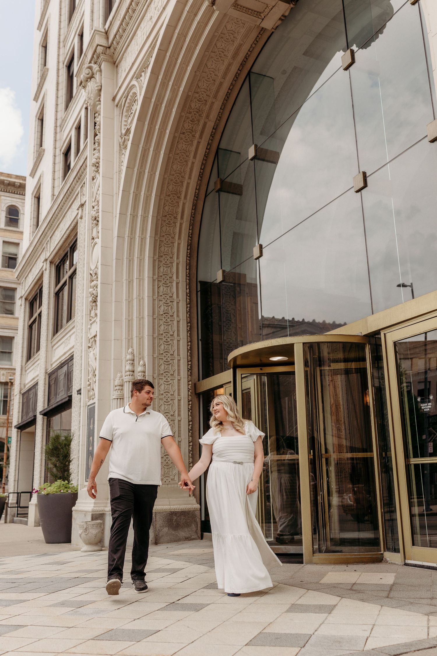 A couple walks together under ornate stone archways along a grand building exterior with tall windows.