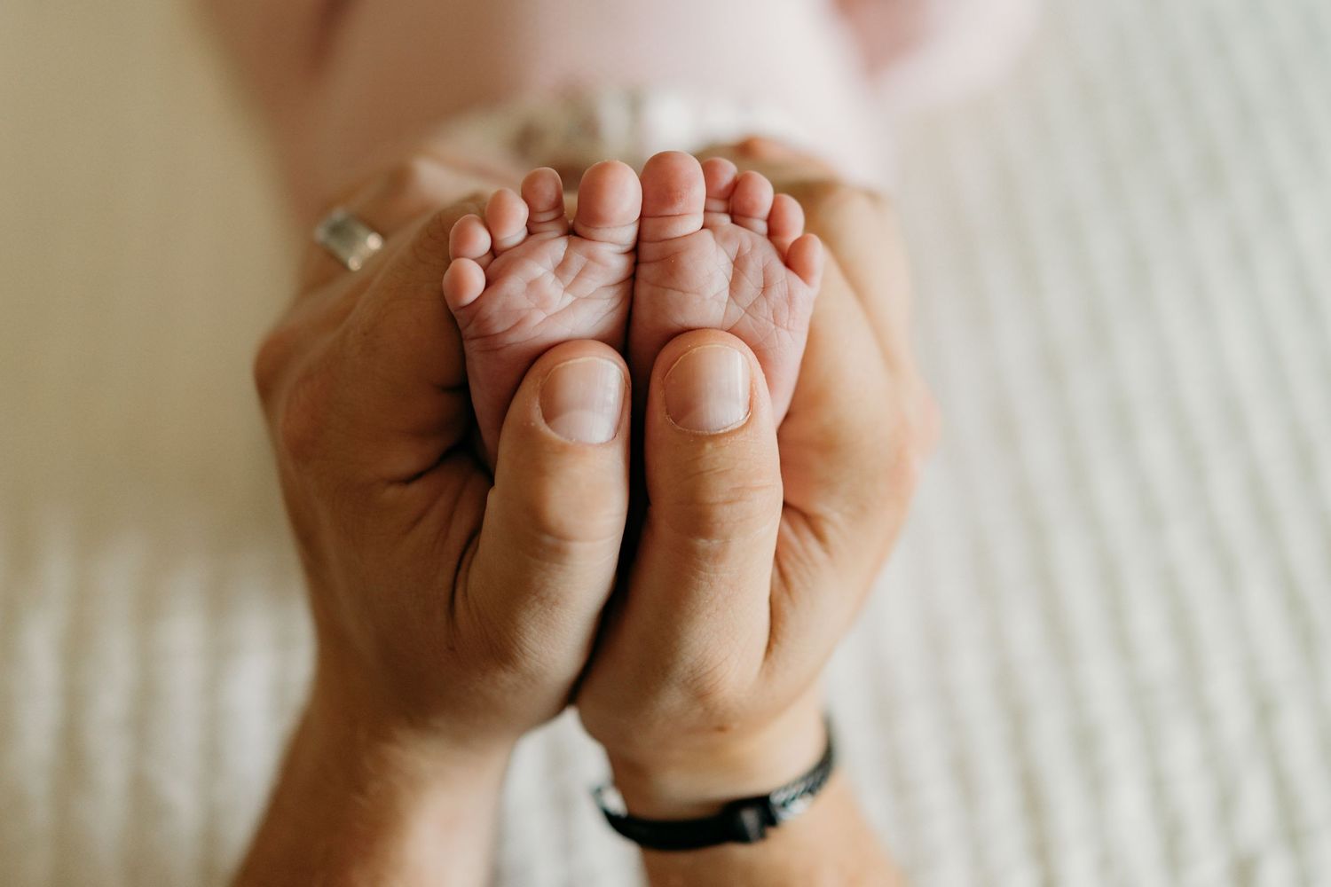 Adult hands gently holding tiny baby feet against white textured background with soft natural lighting.