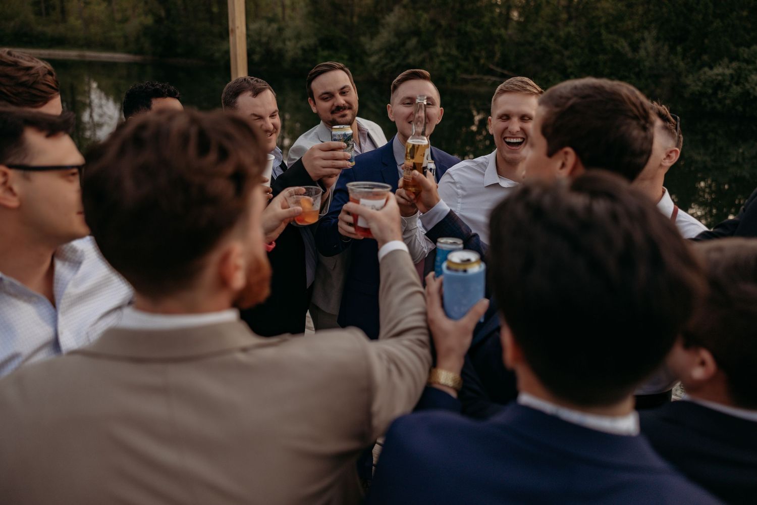 Groomsmen laughing and toasting drinks together outdoors at a wedding celebration.