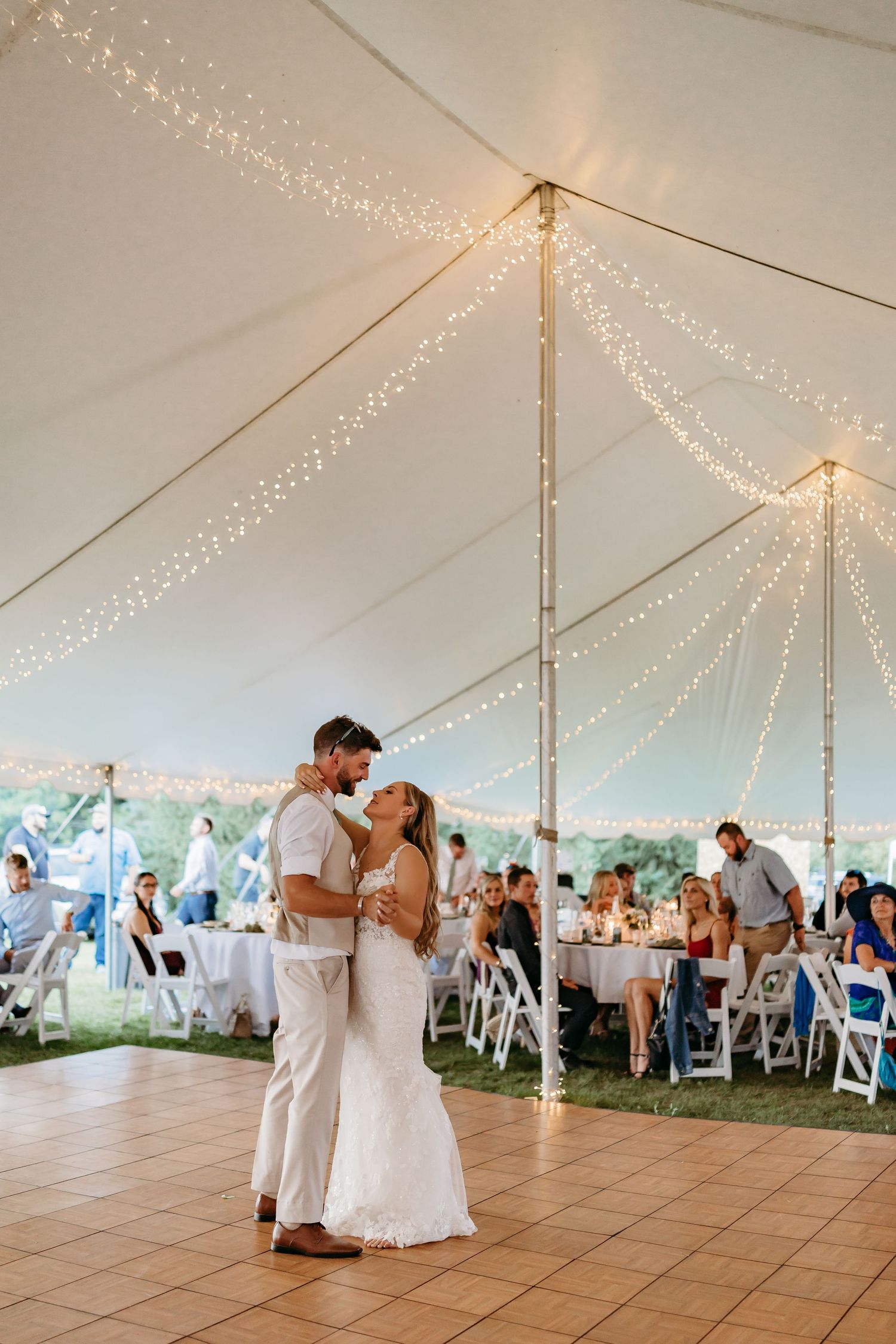 Wedding couple shares their first dance under a white tent decorated with twinkling string lights at an outdoor reception.