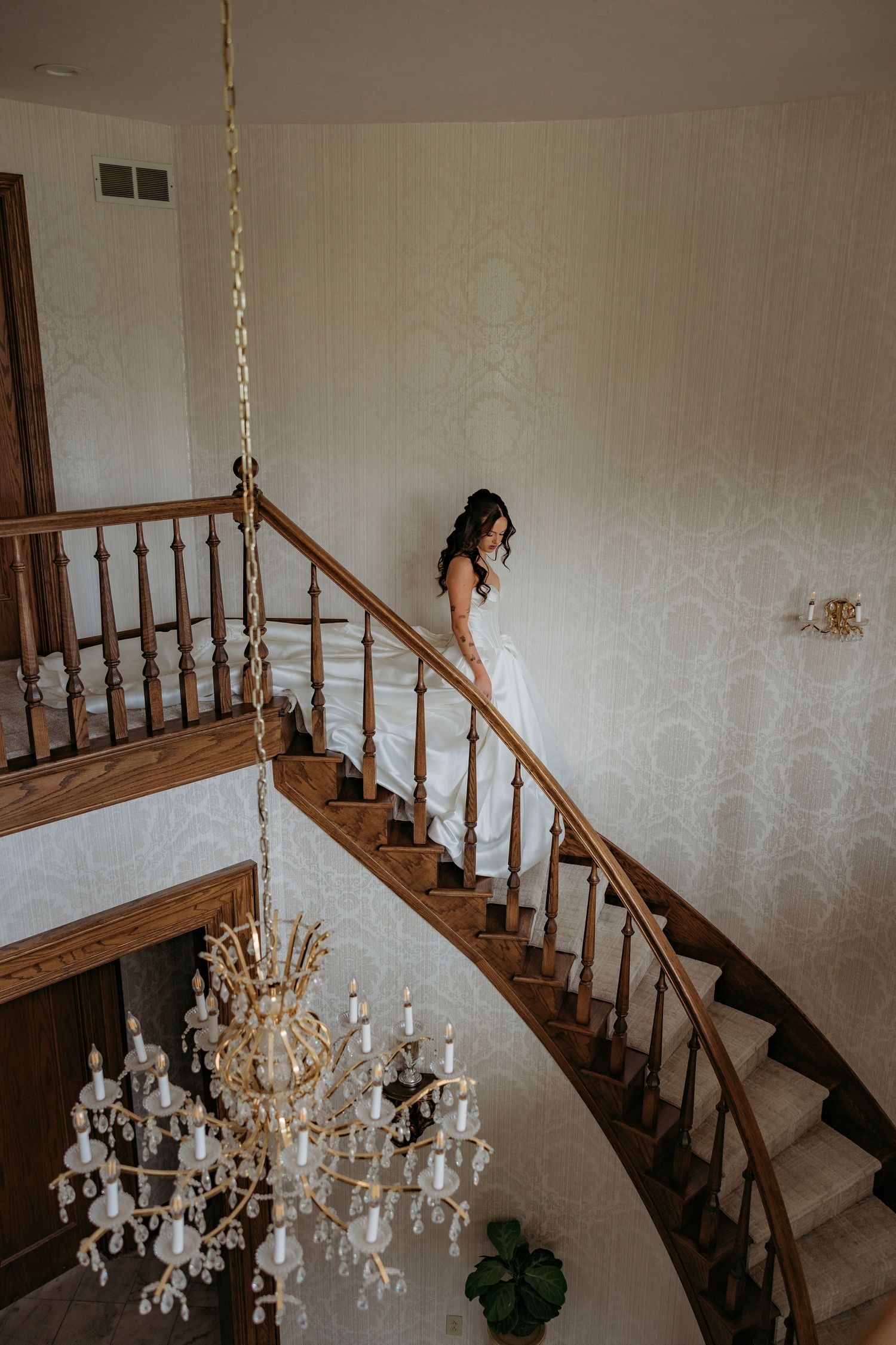 Bride in white gown descending curved wooden staircase with crystal chandelier in elegant venue.