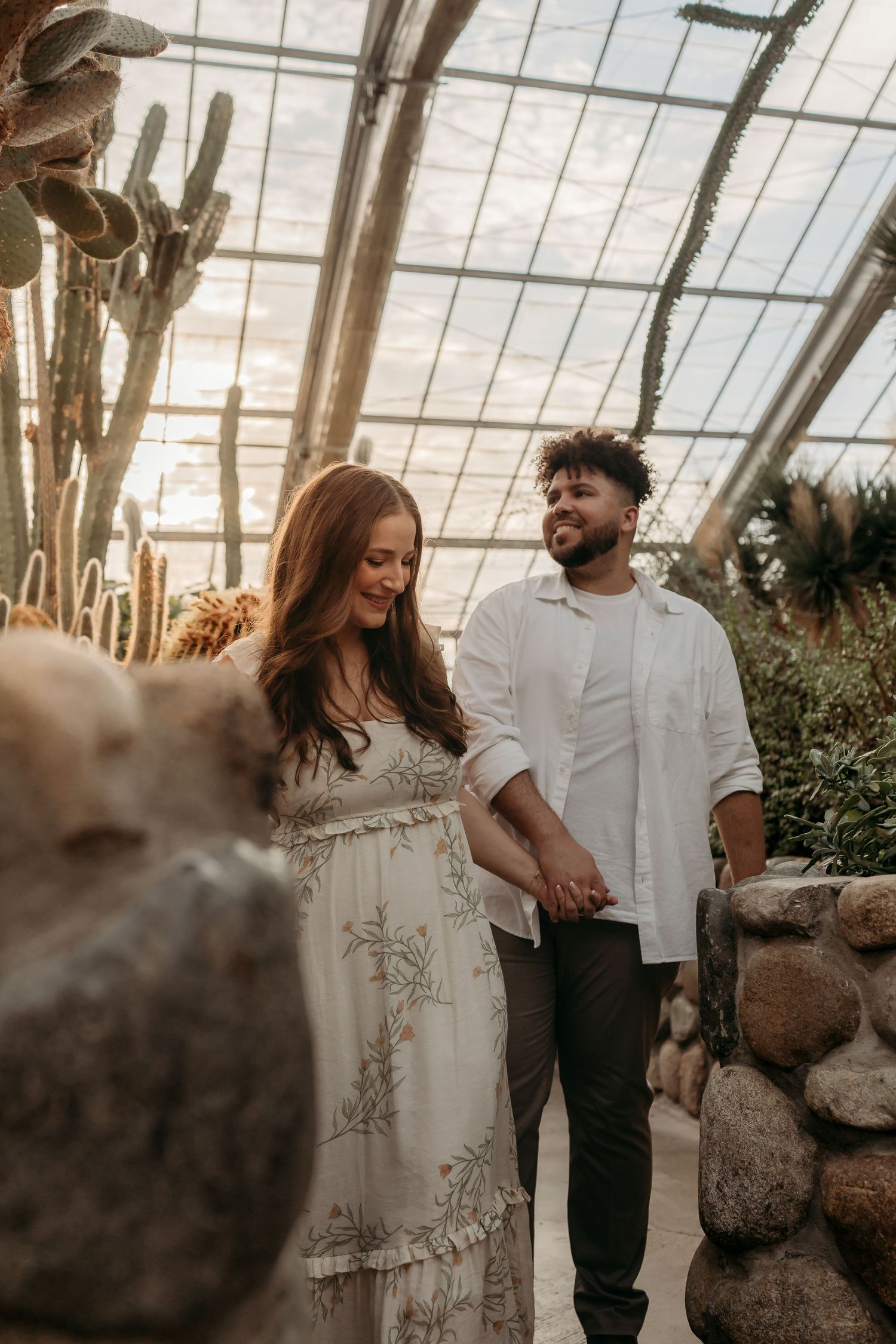 A couple shares an intimate moment together in a sunlit greenhouse filled with tropical plants and cacti.