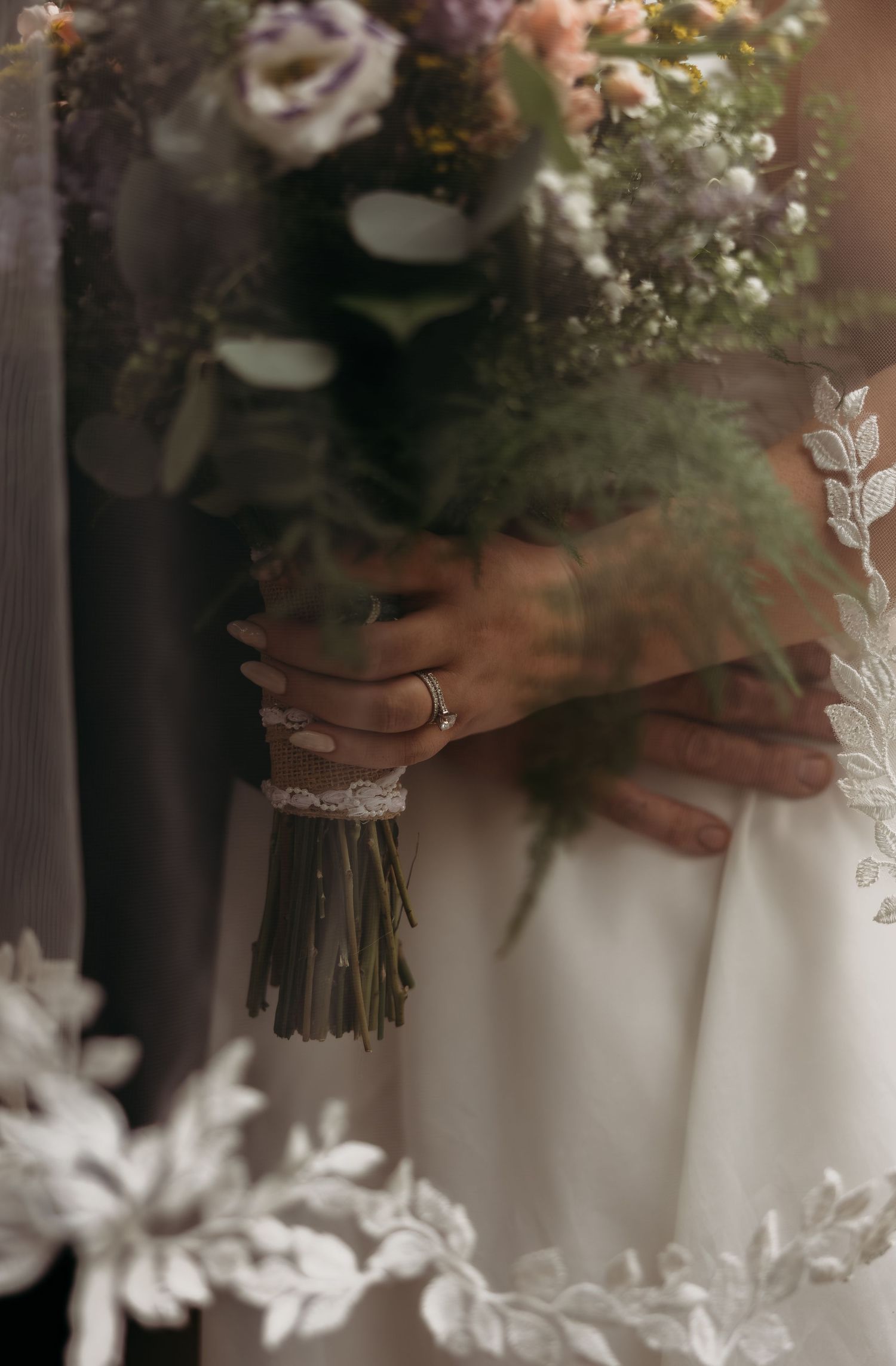 Bride holding a lush wedding bouquet with purple and white flowers, wearing a lace gown and silver ring.