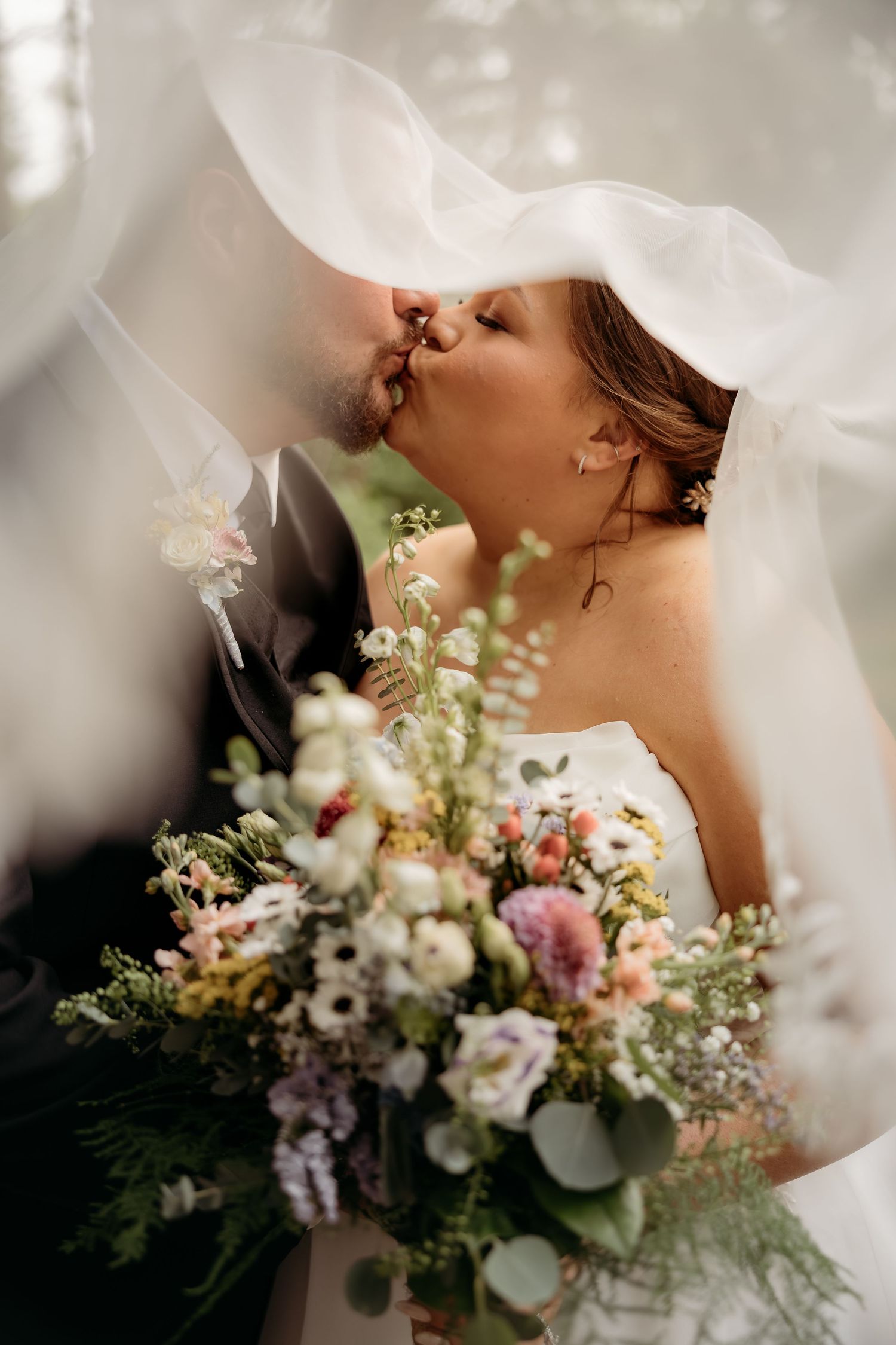 Bride and groom share a romantic kiss under a flowing white veil, bride holding a lush wildflower bouquet.
