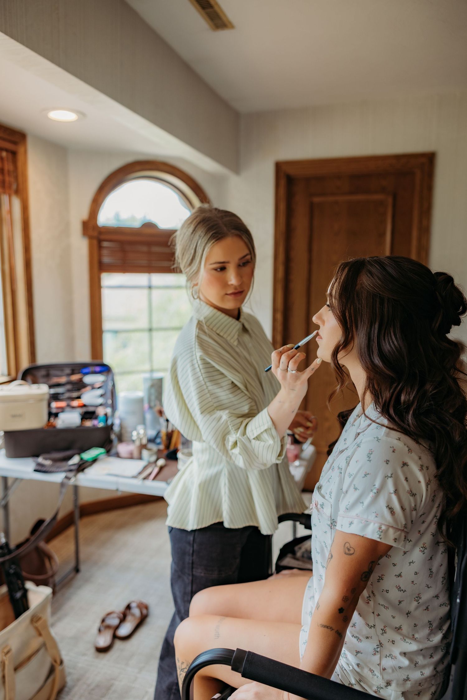 Makeup artist applying lipstick to a bride during wedding day preparations in a bright dressing room.