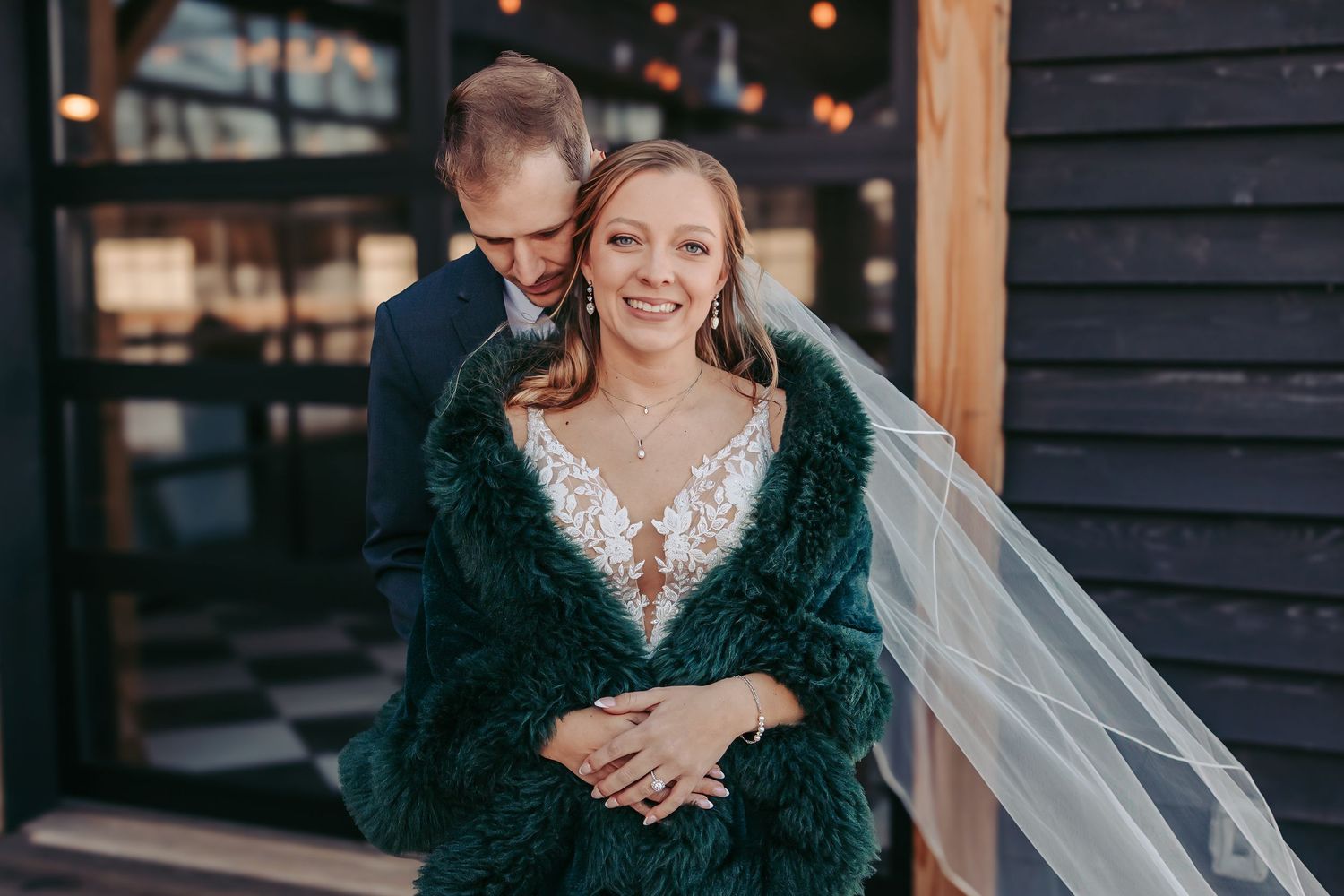 A wedding photo outside a dark building showing a couple embracing with the bride wearing a dark fur shawl and veil.