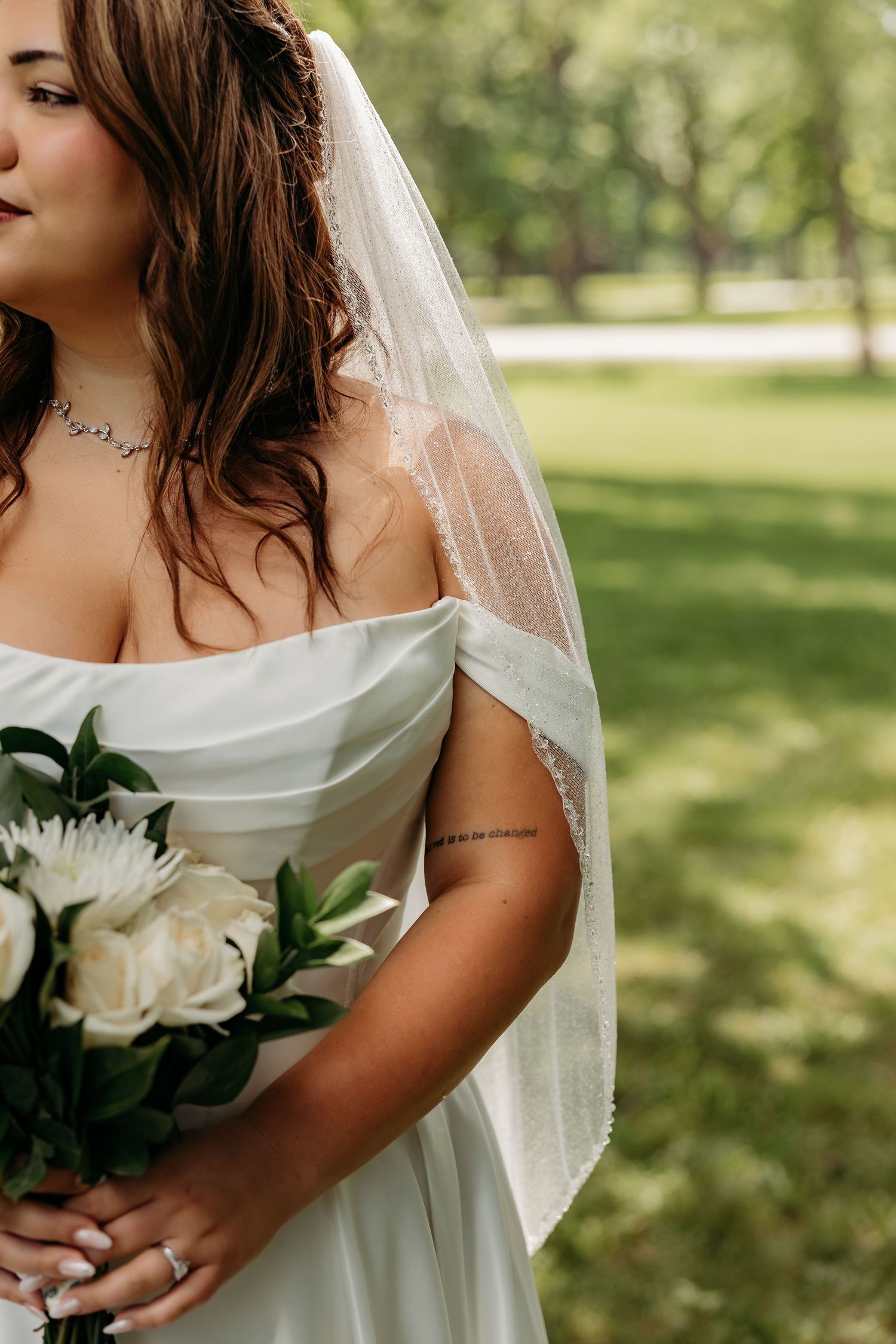 Bride in off-shoulder white gown holding bouquet of white roses, wearing veil and neck tattoo, outdoors.