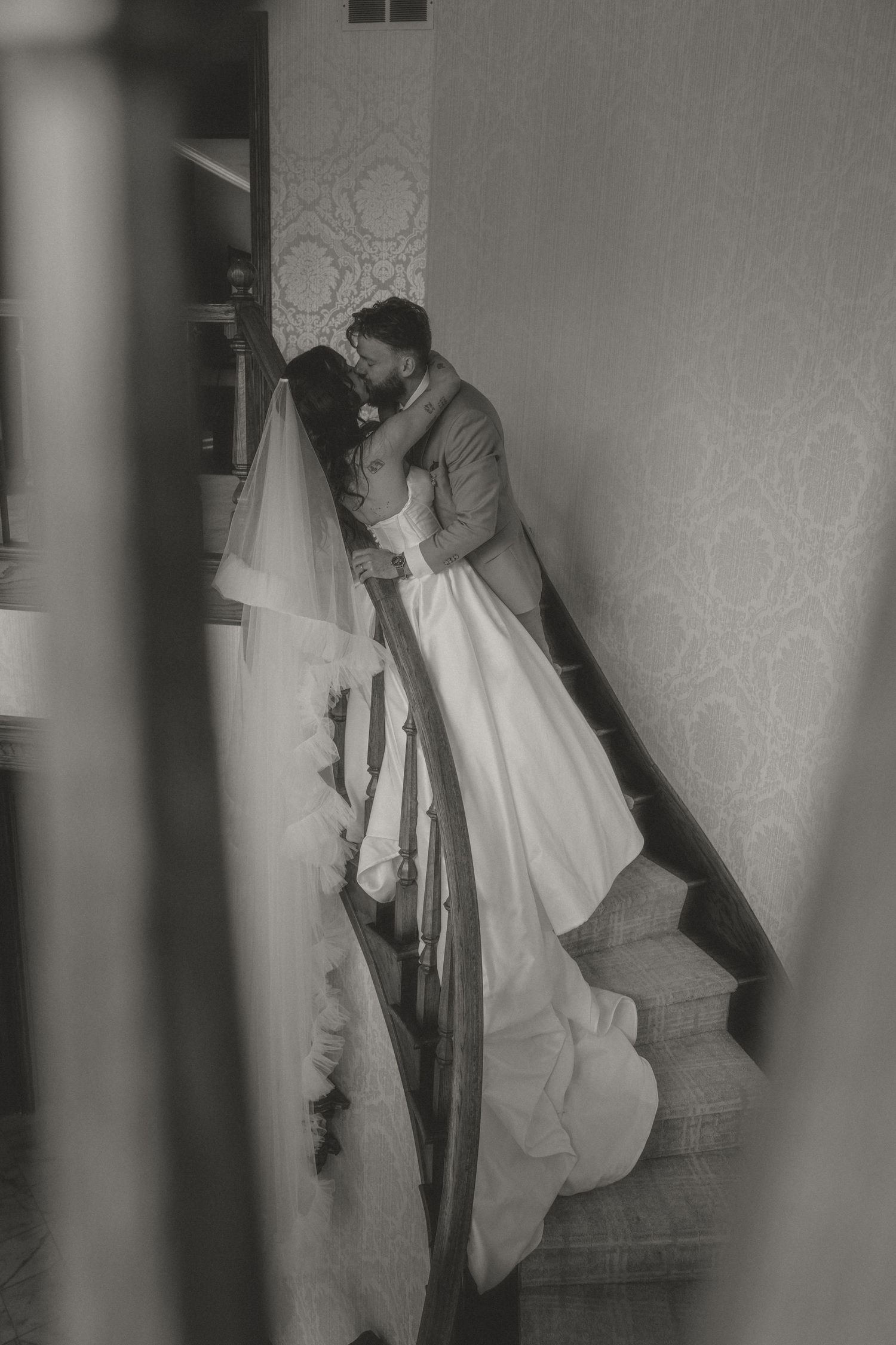 Black and white photo of a bride and groom sharing a romantic kiss on an elegant staircase in a wedding venue.