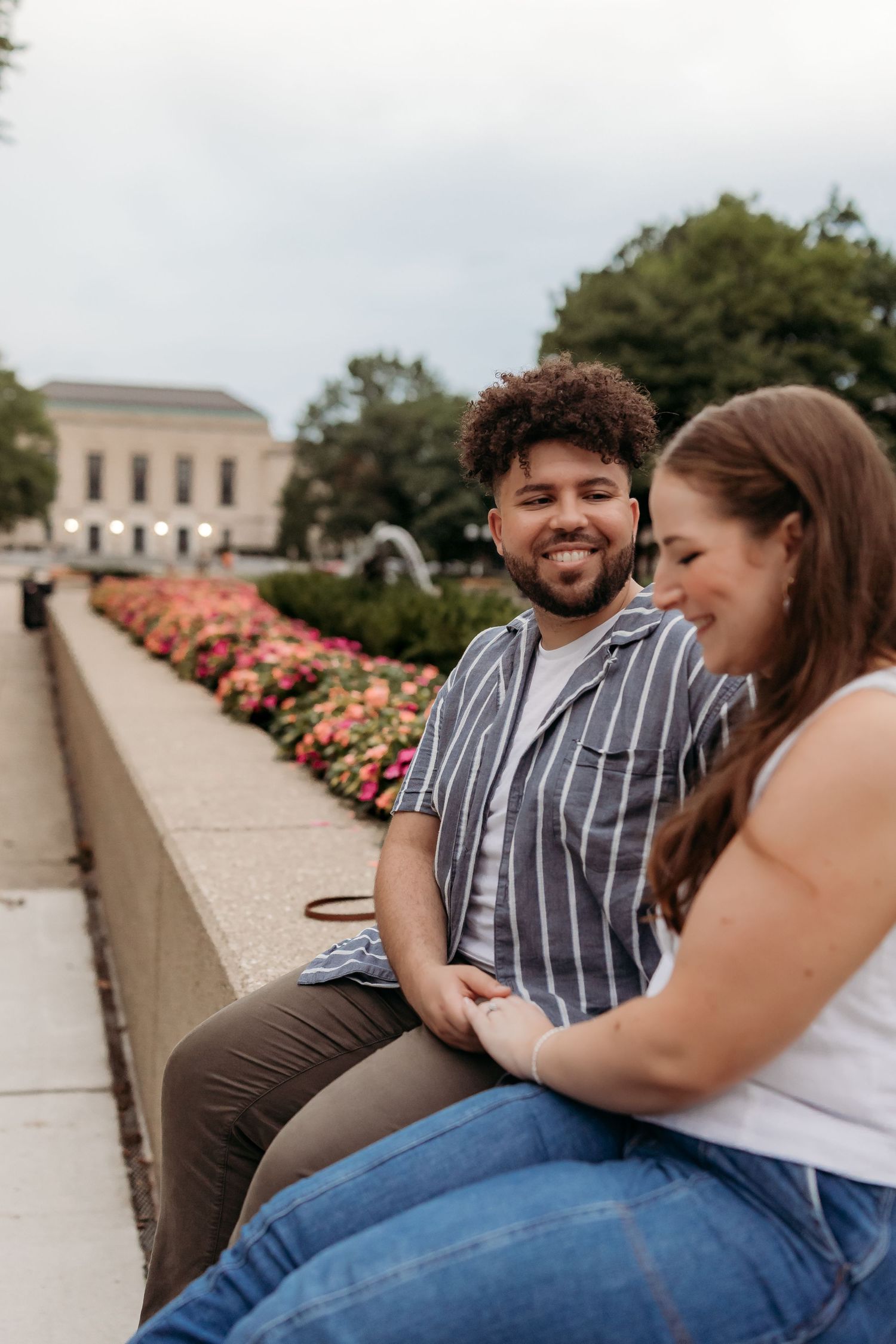 A couple sits together on a concrete wall lined with pink flowers in a garden setting.