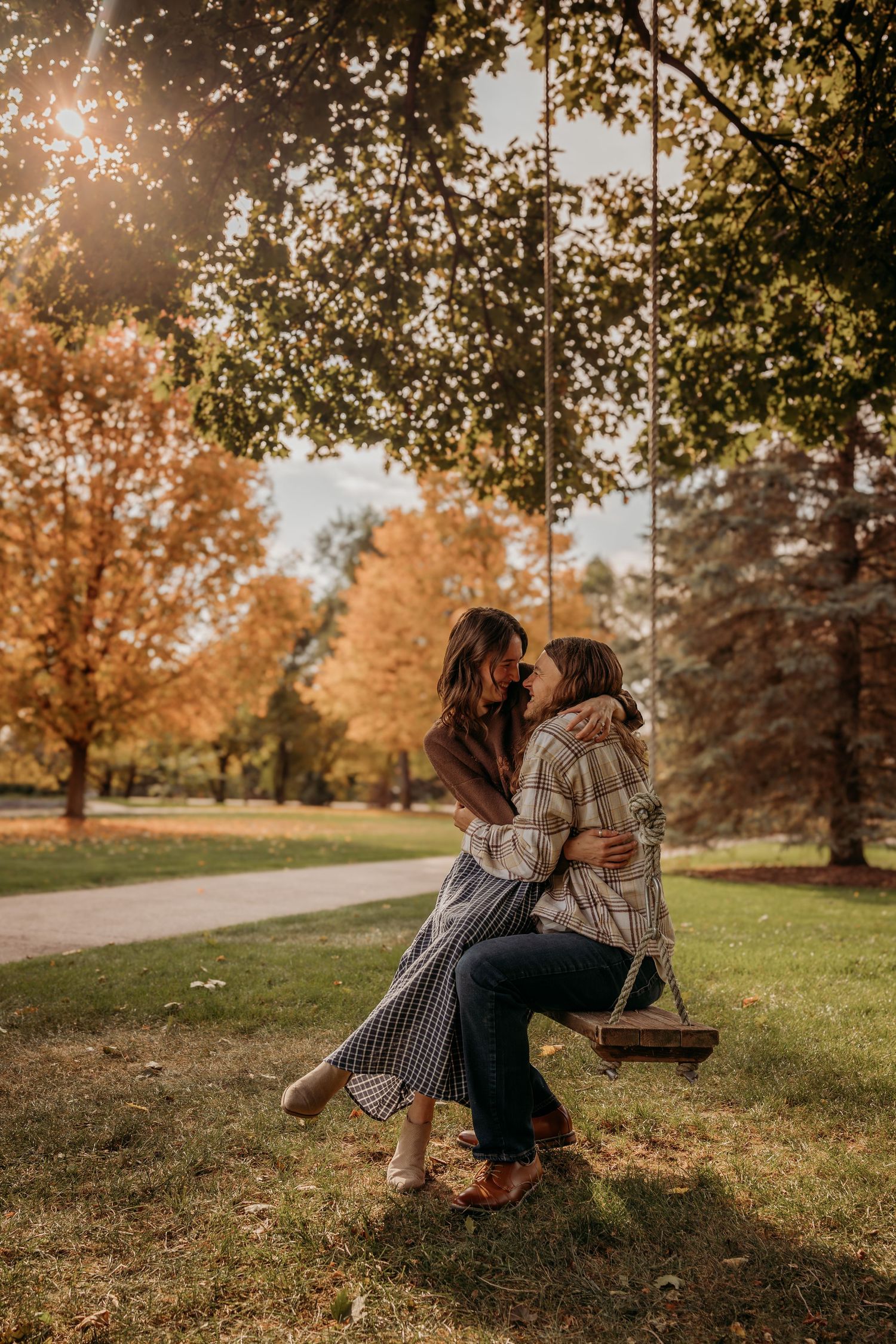 A couple shares a romantic moment on a wooden swing in a sunlit autumn park with golden foliage in the background.