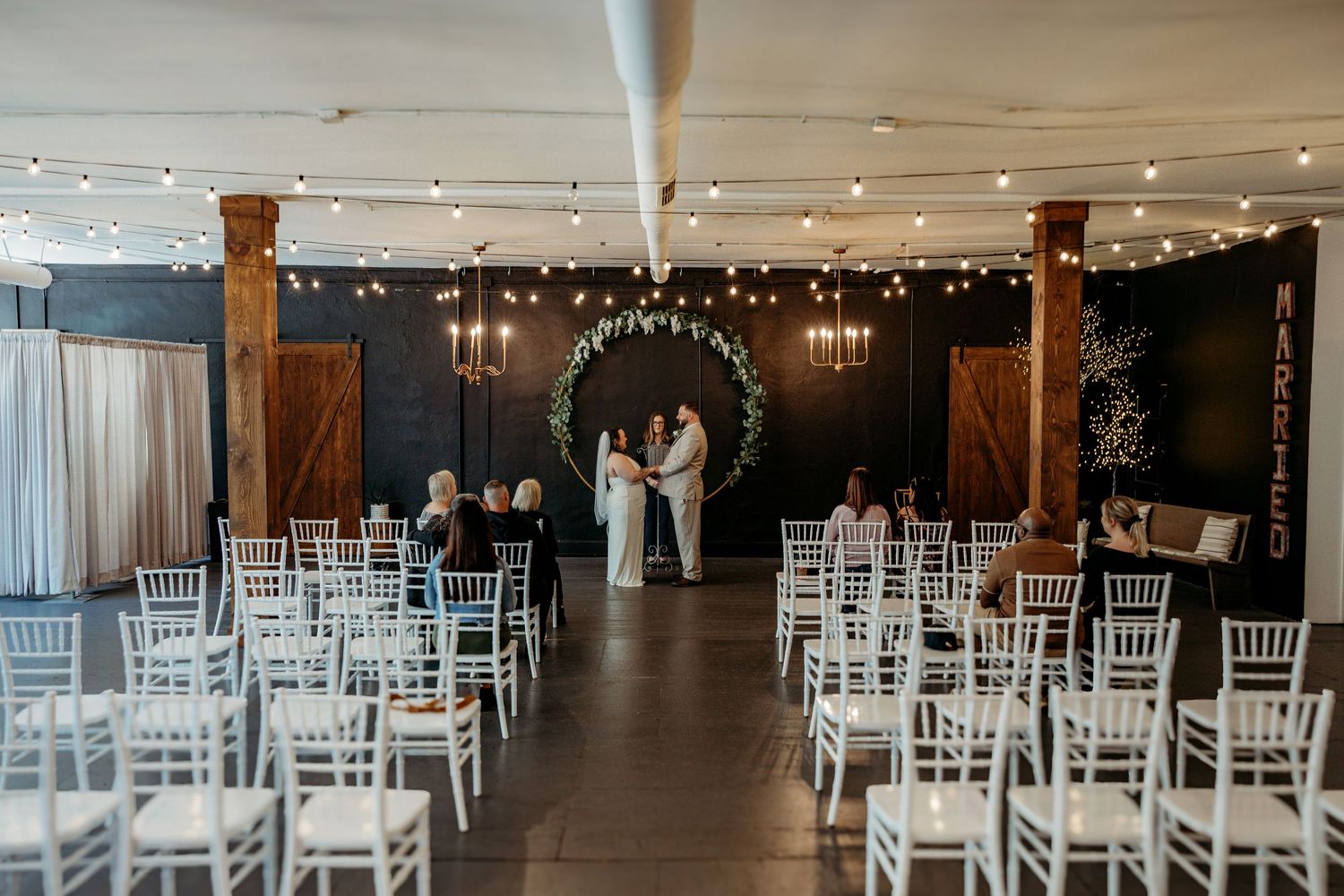 Empty wedding venue decorated with white Chiavari chairs and string lights hanging from wooden beams creates an elegant atmosphere.