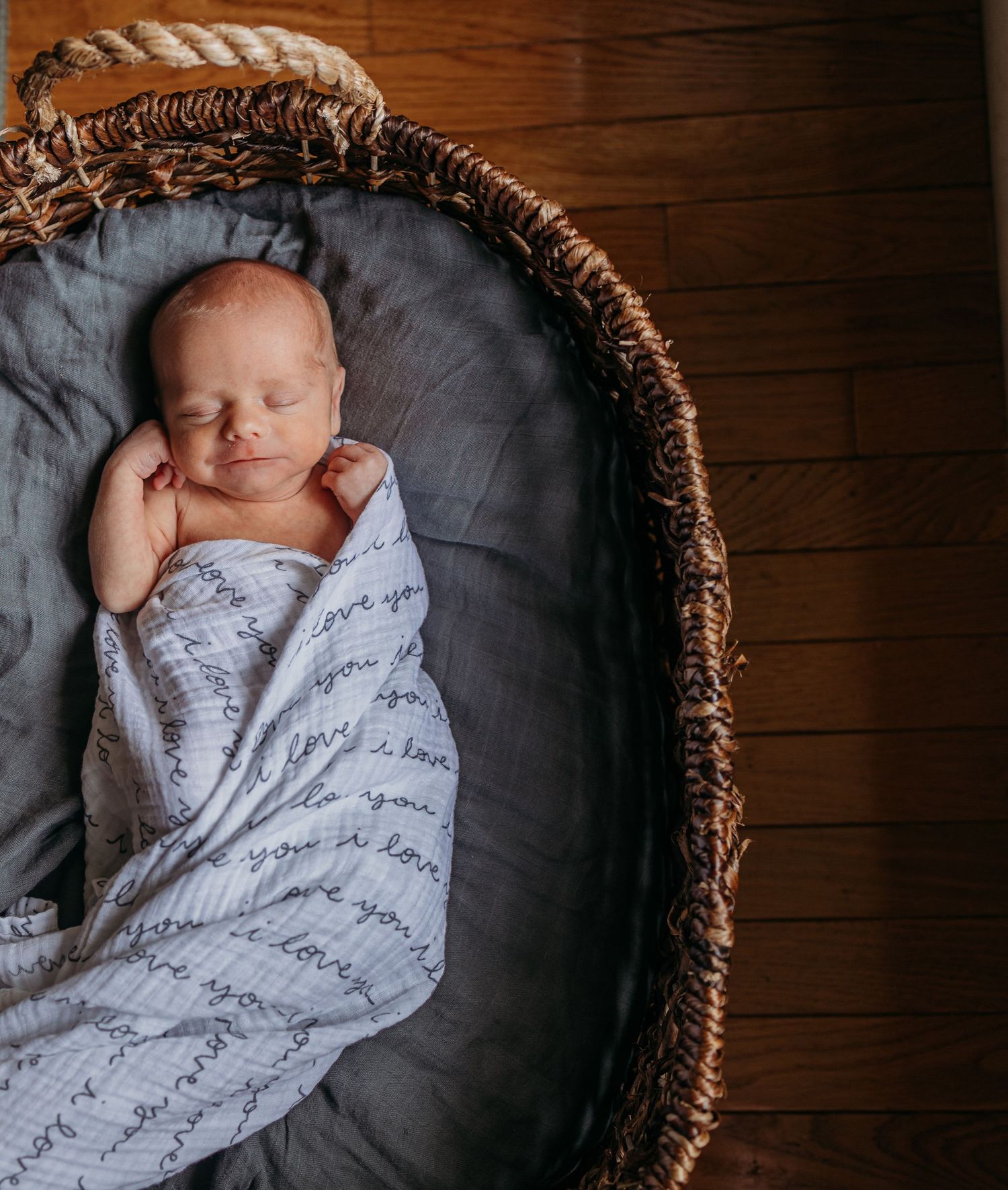 Peaceful sleeping newborn wrapped in a white patterned swaddle blanket rests in a cozy woven bassinet.