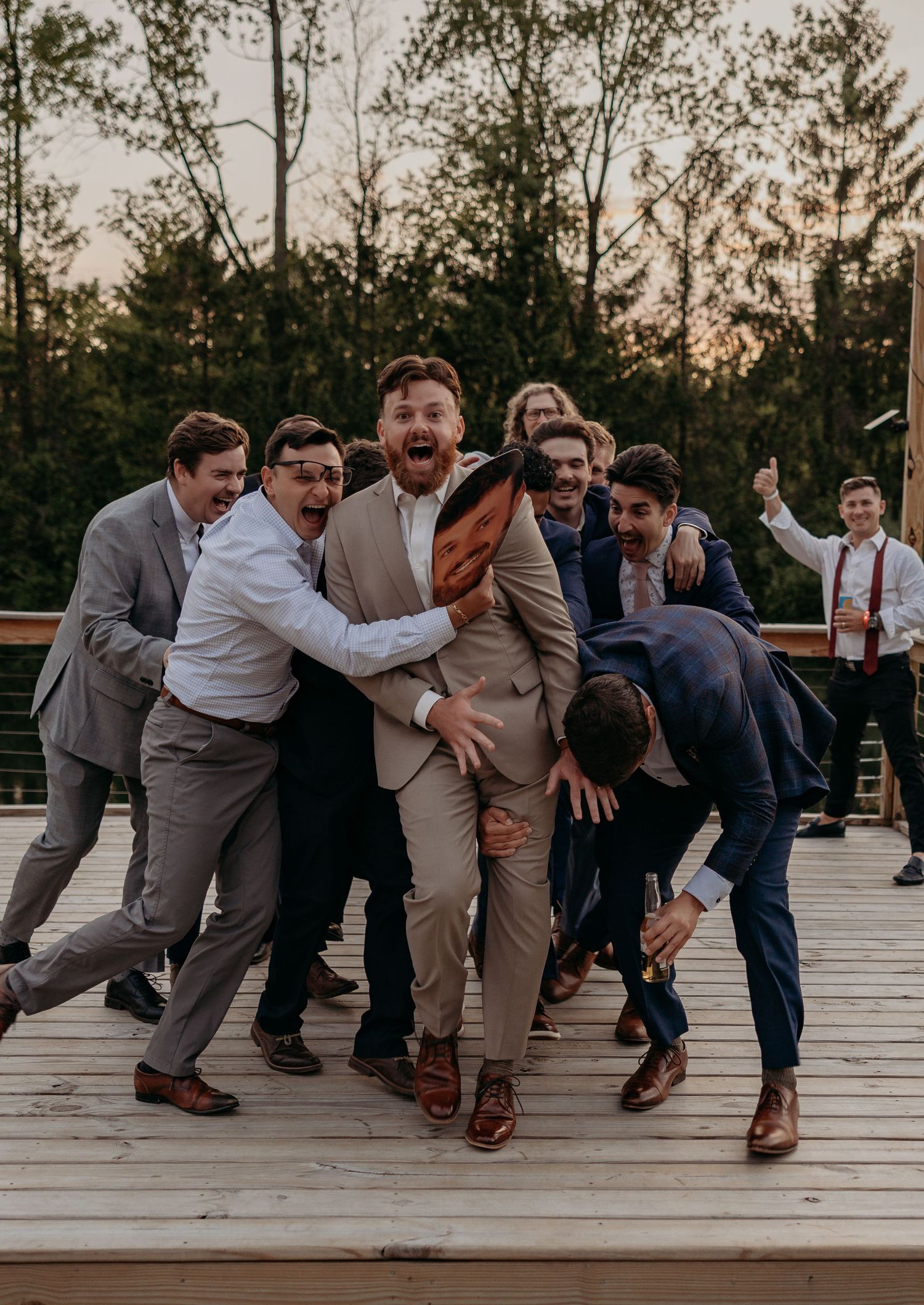 Excited groom in tan suit celebrates with groomsmen on wooden deck at outdoor wedding reception.