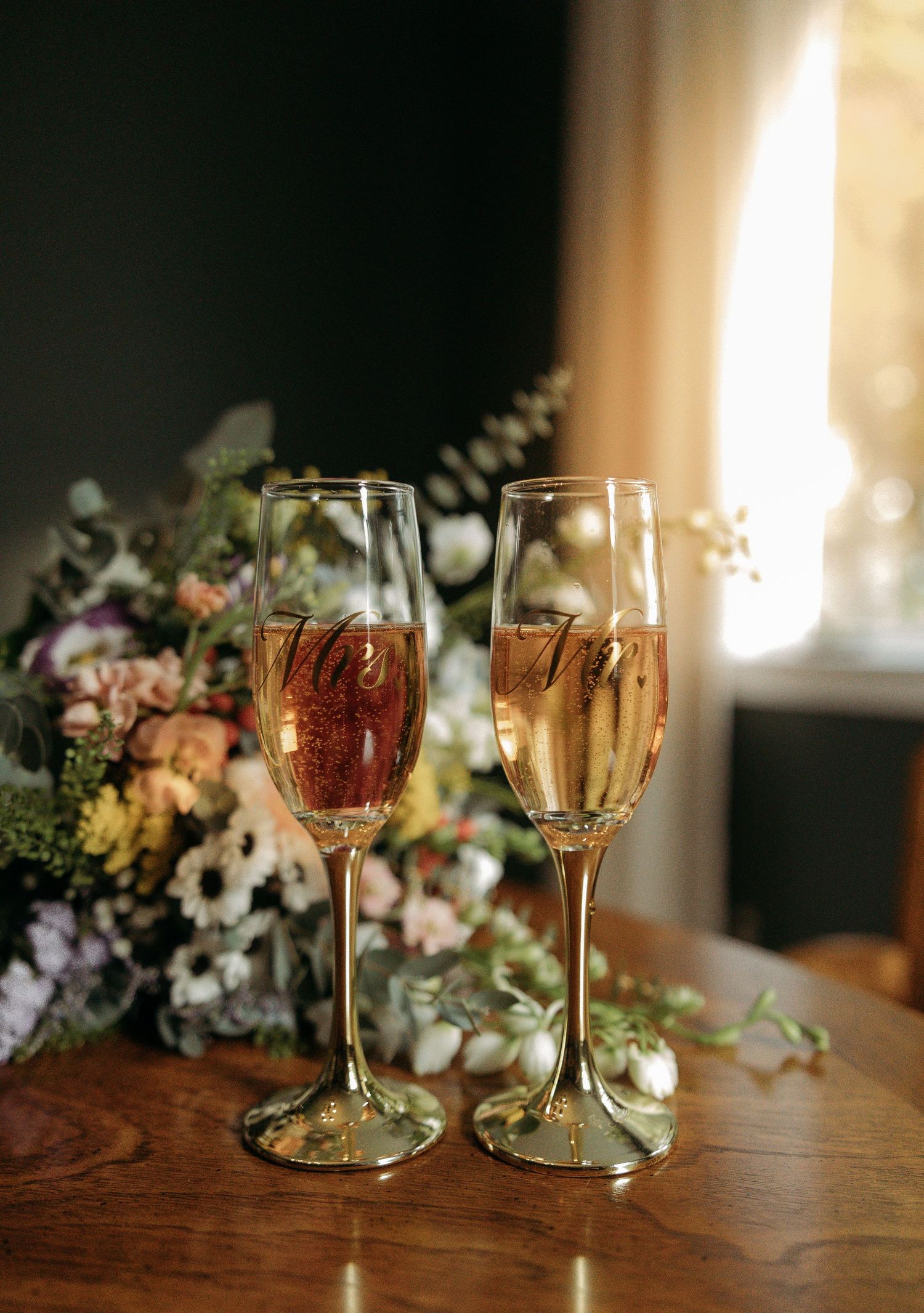 Two gold champagne flutes with Mr. and Mrs. engravings beside a floral bouquet on a wooden table.