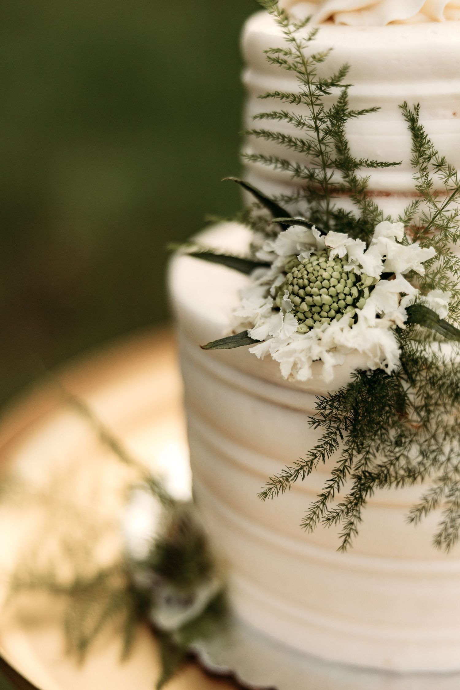 Close-up view of a white wedding cake decorated with delicate fern leaves and a single white scabiosa flower on a gold surface.