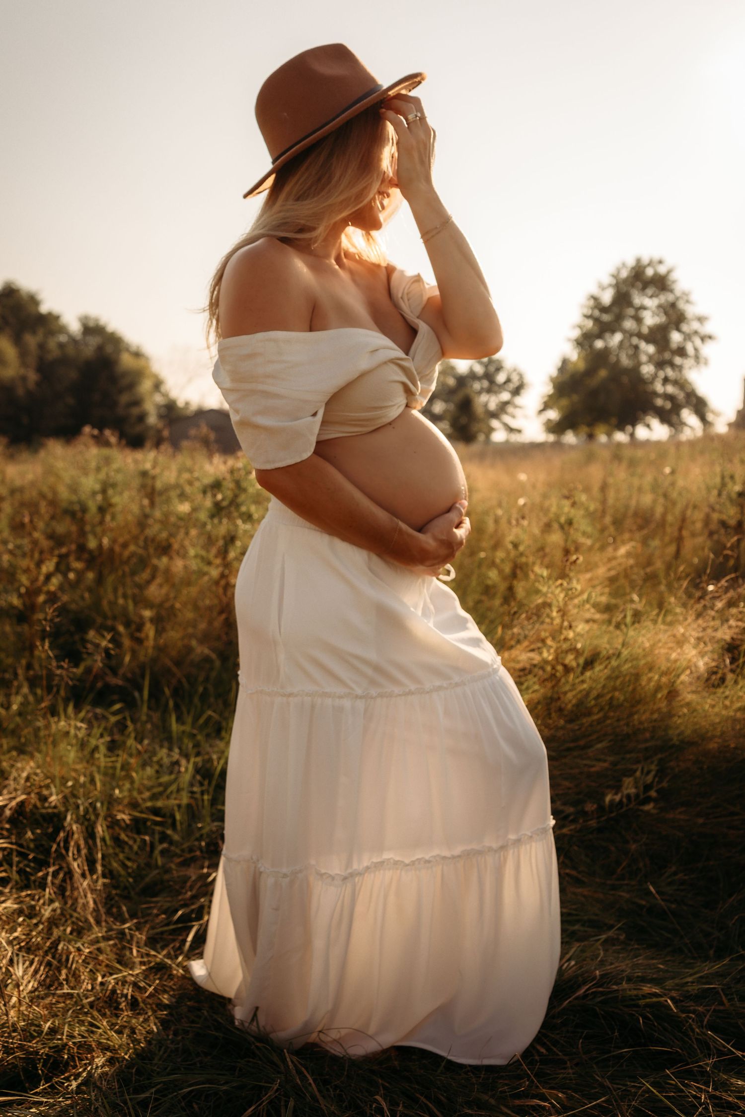 A pregnant woman in a white dress and brown hat poses in a sunlit field at golden hour, holding her belly.