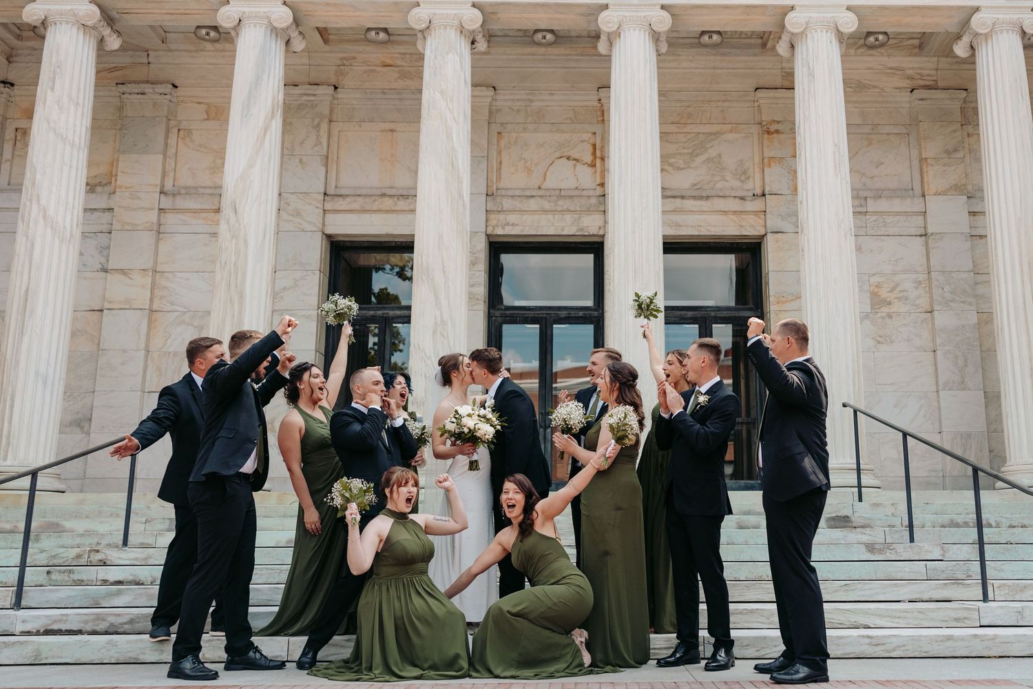 Wedding party in formal attire poses joyfully on grand stone steps with white columns of classical building.