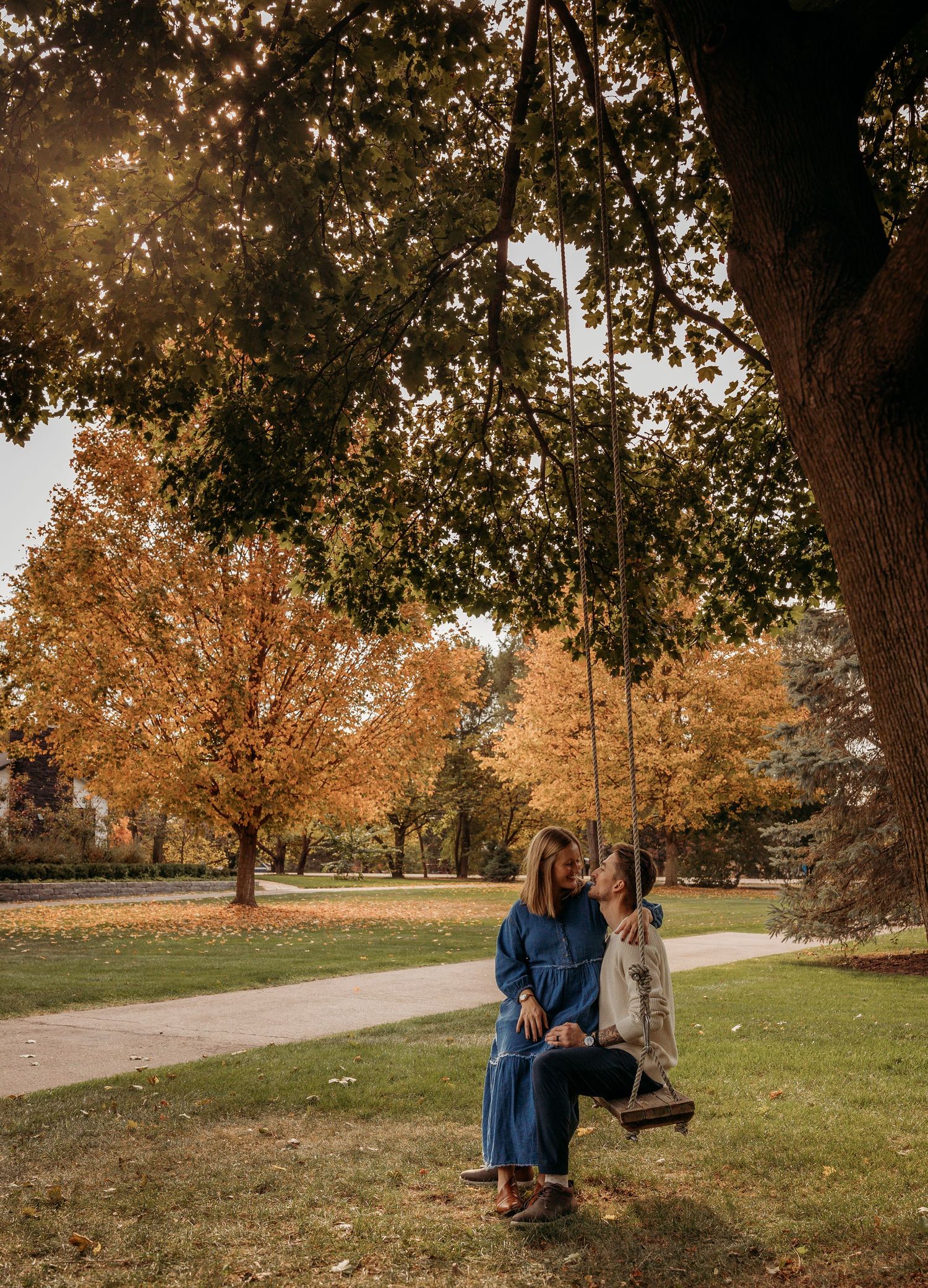 A person in denim sits on a wooden swing under autumn trees in a peaceful park setting.