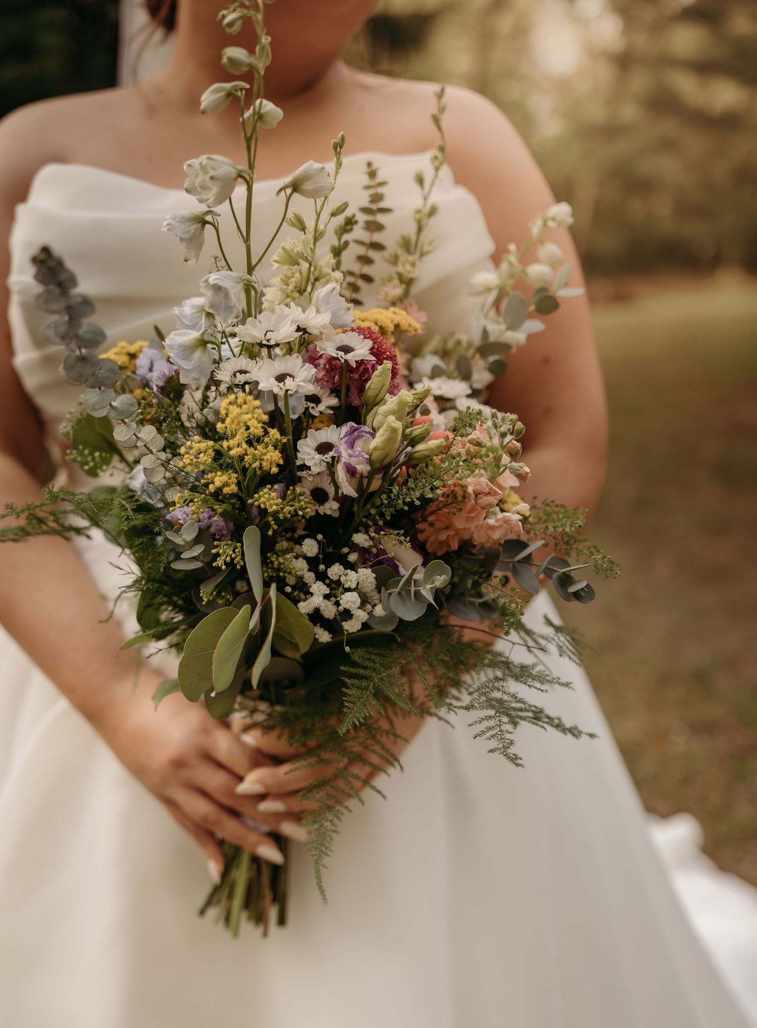 Bride in white strapless gown holding a wildflower bouquet with purple, yellow, and pink blooms and lush greenery.