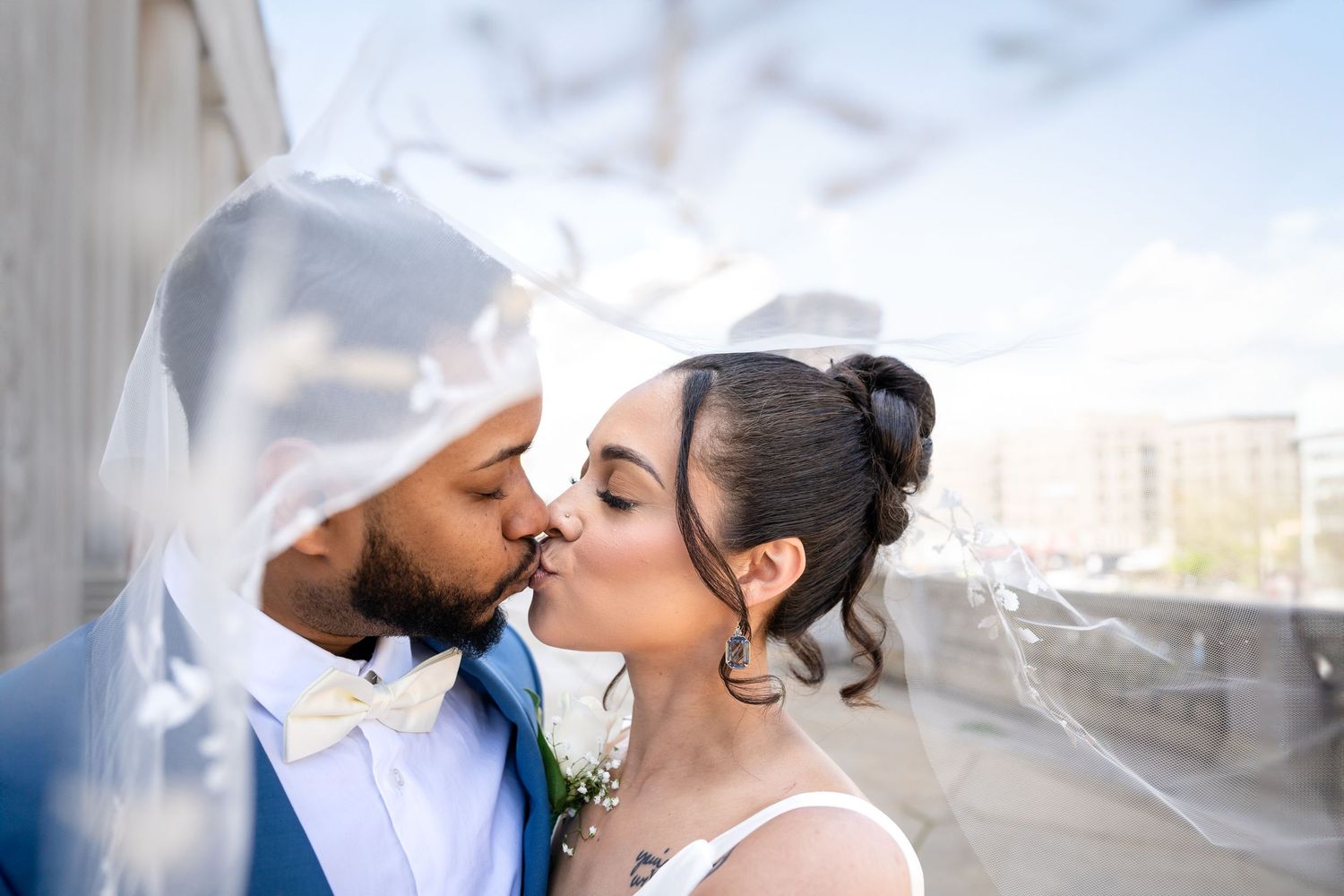 A bride and groom share a kiss under a sheer wedding veil, with a city skyline softly blurred behind them.