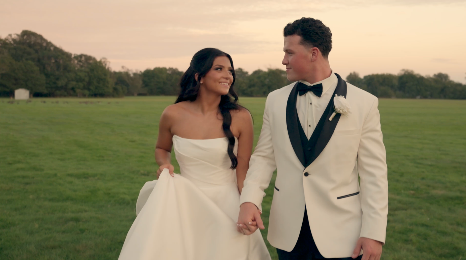 Bride in a strapless white gown and groom in a cream tuxedo jacket hold hands while walking across a green field at sunset.