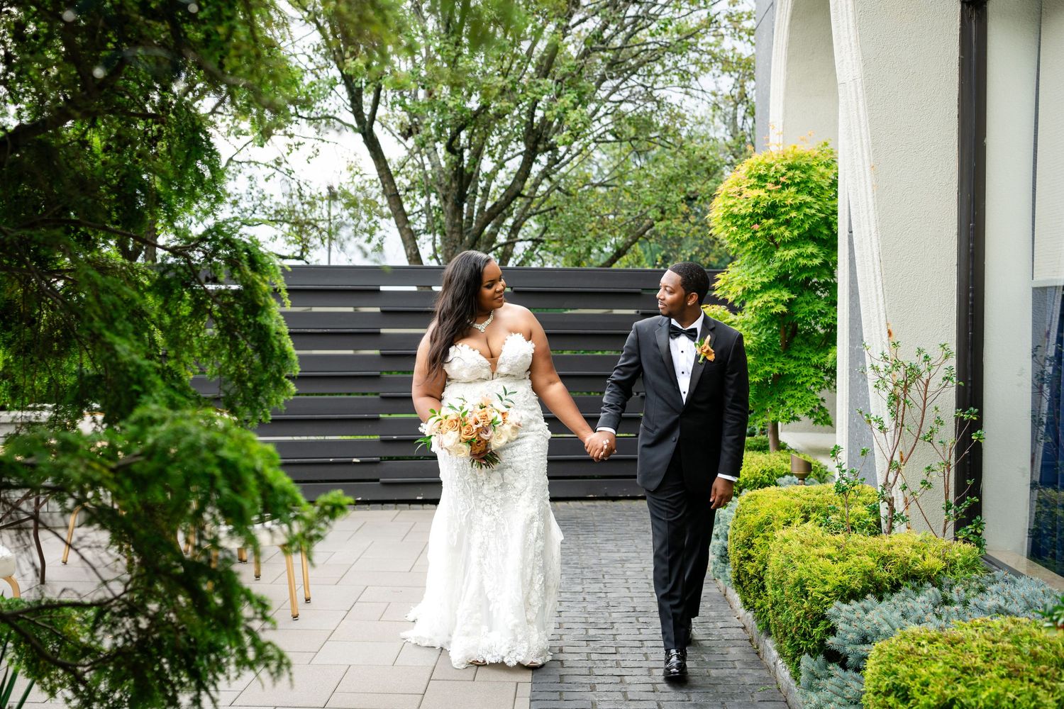 Bride in a white lace gown holding a bouquet walks hand in hand with a groom in a black tuxedo along a stone path lined with green shrubs.