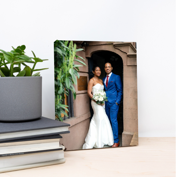 Wedding photo print of a bride in a white gown holding a bouquet beside a groom in a blue suit, displayed on a shelf with books and a plant.