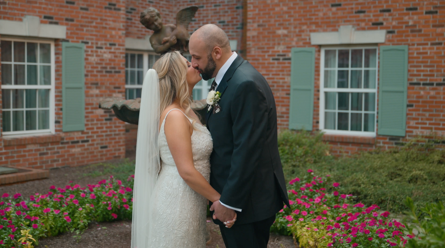 Bride and groom share a kiss in front of a brick building with green shutters, surrounded by colorful flower beds.