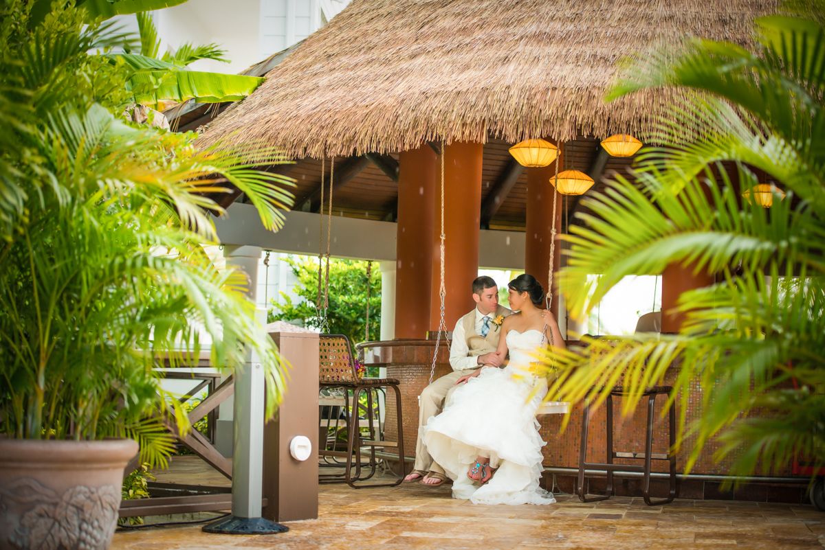 Bride in white gown and groom sit close together under a thatched-roof tropical pavilion surrounded by lush green palm leaves.