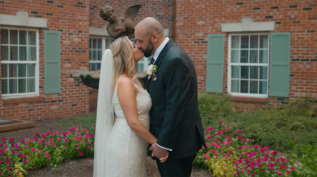A couple shares an intimate moment in front of a brick building surrounded by pink flowers and landscaping.