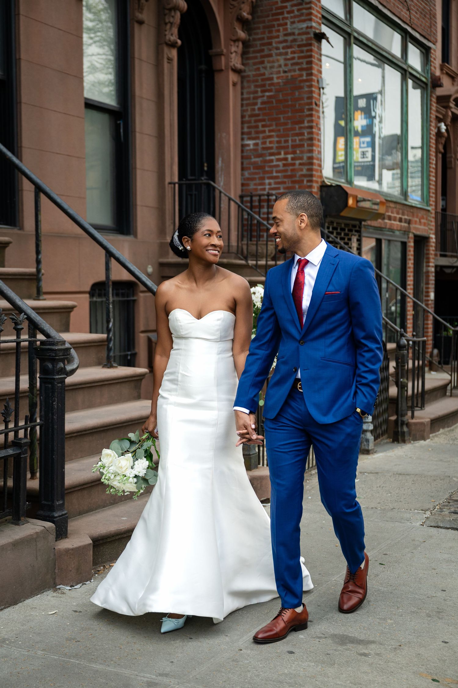 Bride in white strapless gown holding a bouquet walks hand in hand with groom in blue suit past brownstone stairs.