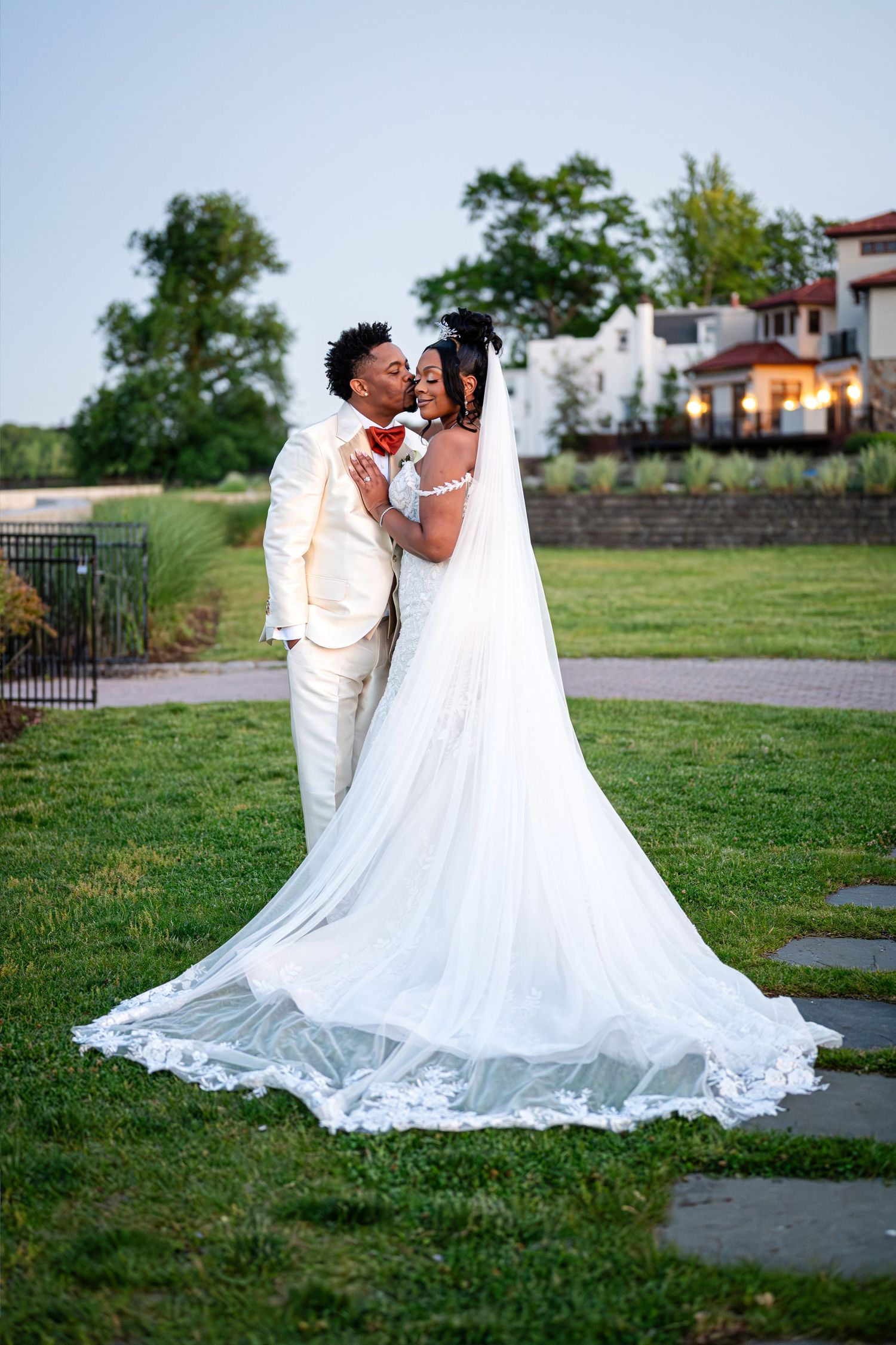 Bride in a flowing white gown with a long lace-trimmed veil kisses her groom in a cream suit on a manicured lawn at an outdoor wedding.