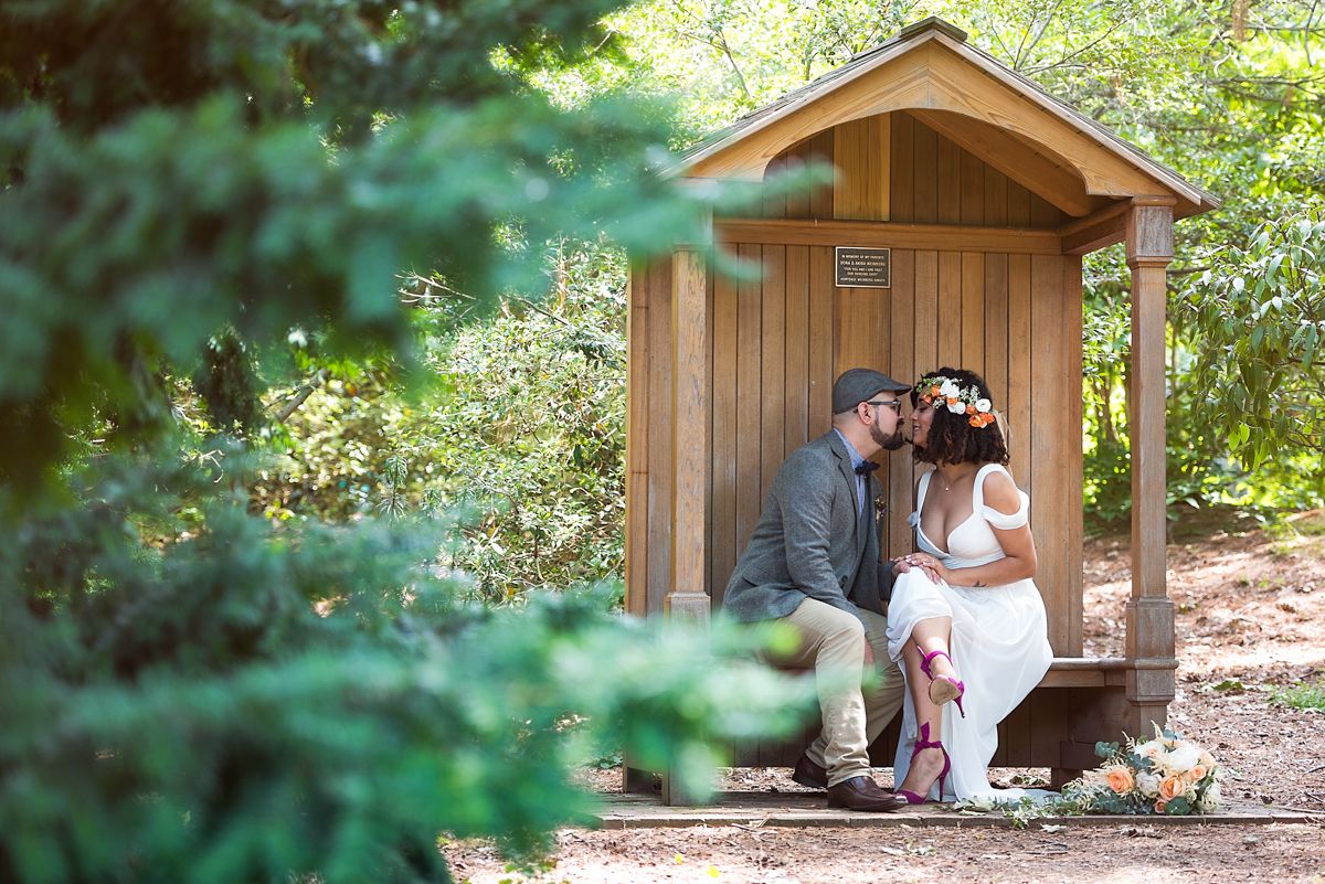 Couple in wedding attire sharing a kiss while seated in a rustic wooden gazebo surrounded by lush green forest foliage.