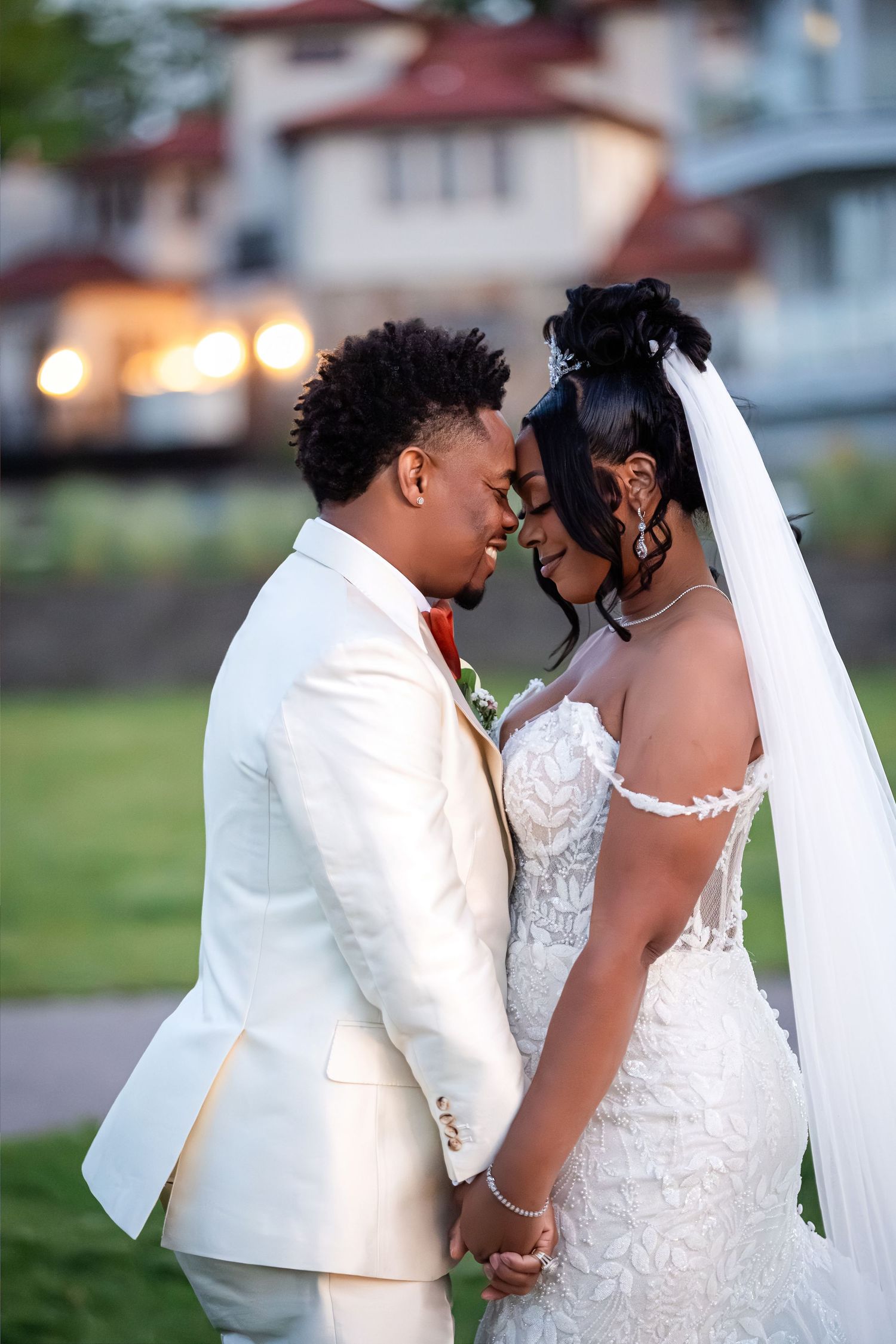 A bride and groom in elegant wedding attire hold hands and touch foreheads tenderly outdoors at dusk.