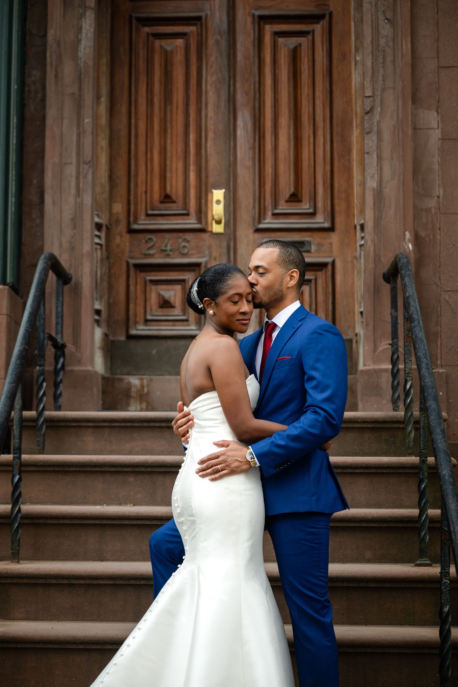 A bride and groom embrace on brownstone steps, she in a white mermaid gown, he in a blue suit with red tie.