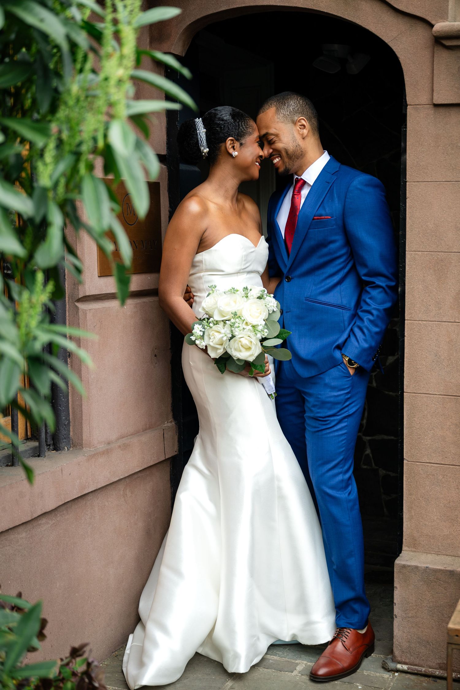 Smiling bride in a white strapless gown holding a white bouquet embraces her groom in a bright blue suit and red tie in a stone doorway.