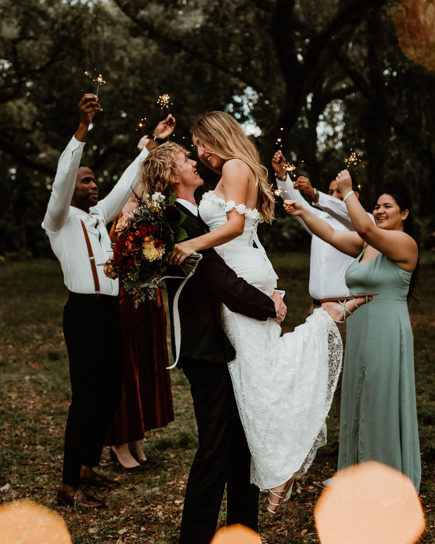 Wedding couple kissing while bridal party celebrates with sparklers outdoors in a wooded setting.