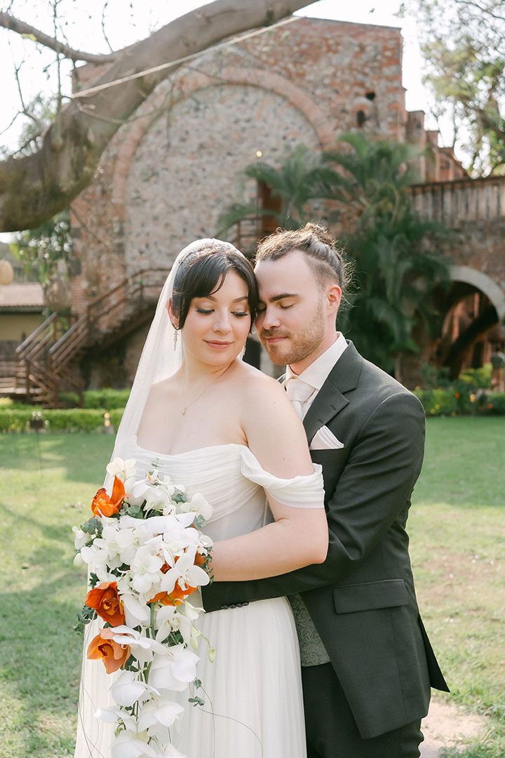 A romantic wedding couple embraces outdoors against a rustic stone wall backdrop with white and orange floral bouquet.