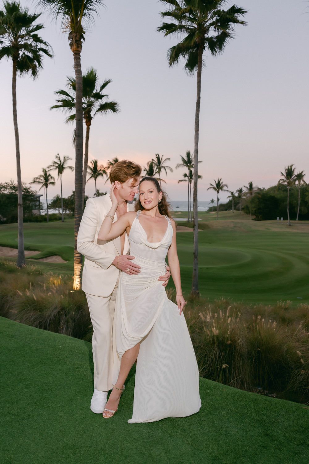 Couple in light-colored attire dances together on a golf course at sunset with palm trees in the background.