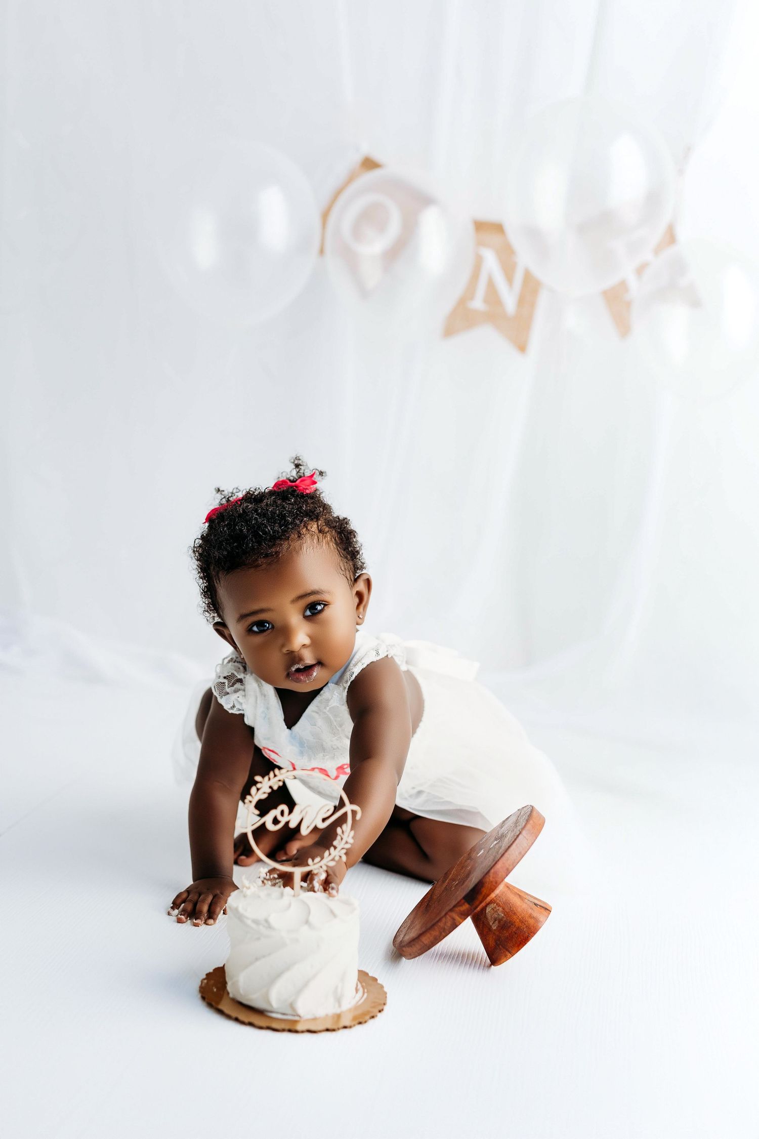A small child in a white outfit sits near a smash cake during a first birthday photoshoot with ONE banner decoration.