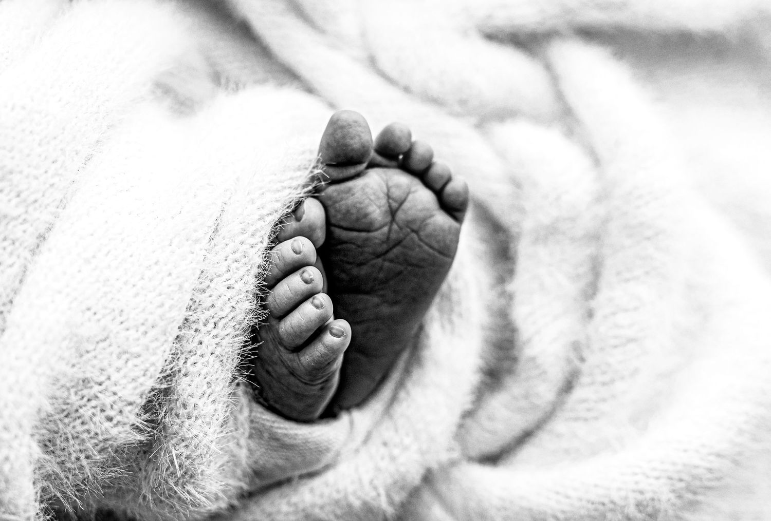 Black and white close-up photo of a newborn baby's tiny foot wrapped in a soft white blanket.
