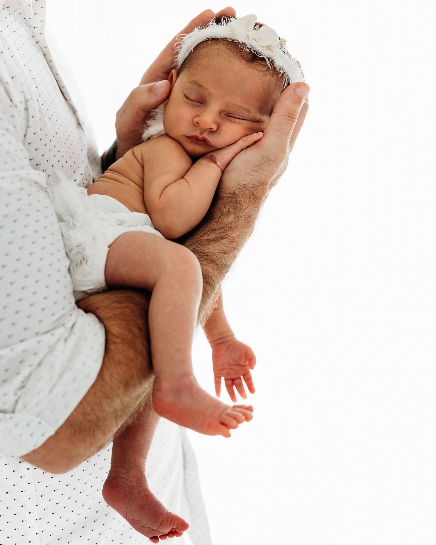 A sleeping newborn baby swaddled in white being gently cradled in strong adult hands against a light background.
