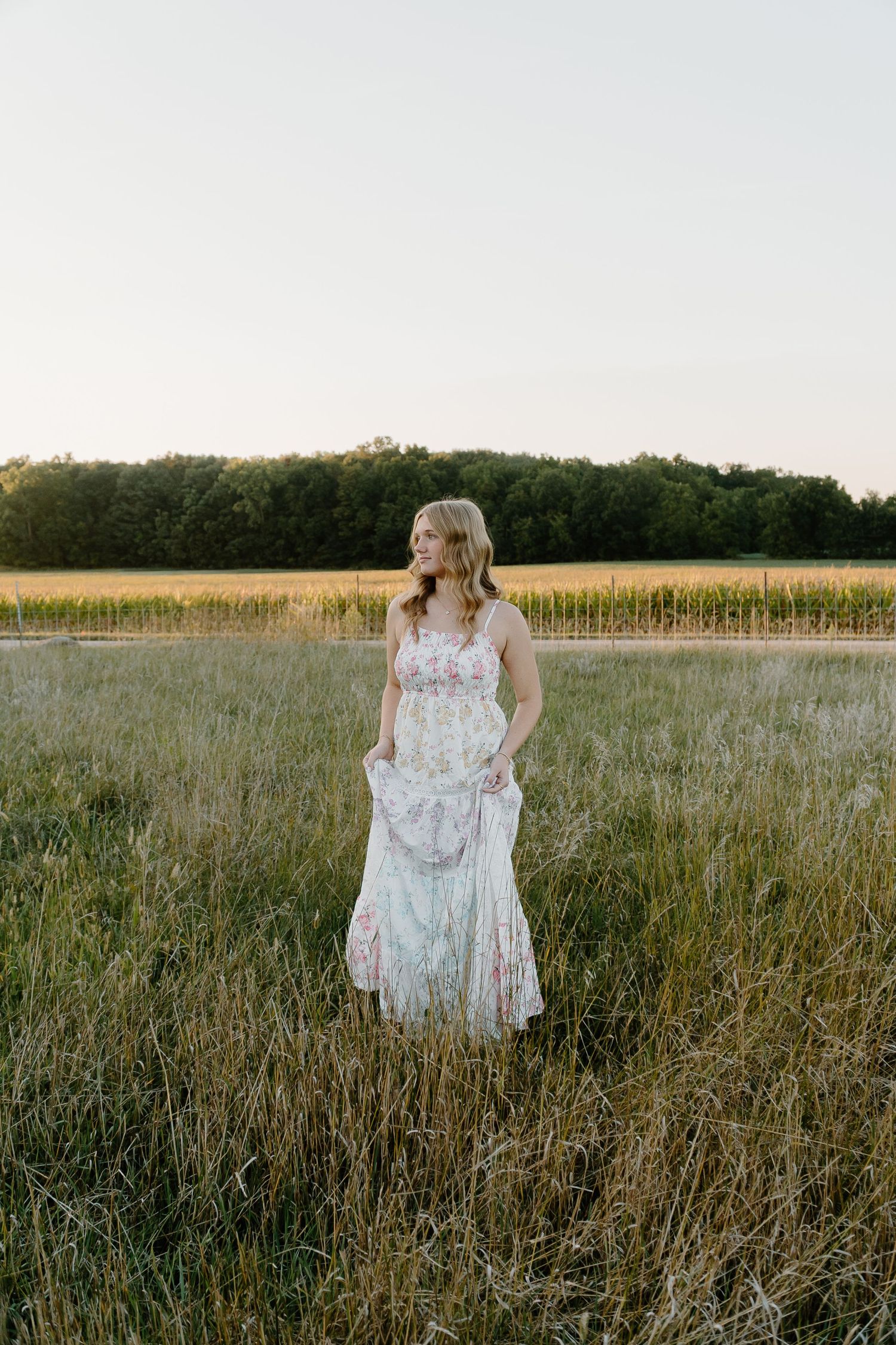 A person in a floral dress walks through a golden field at sunset with trees in the background.
