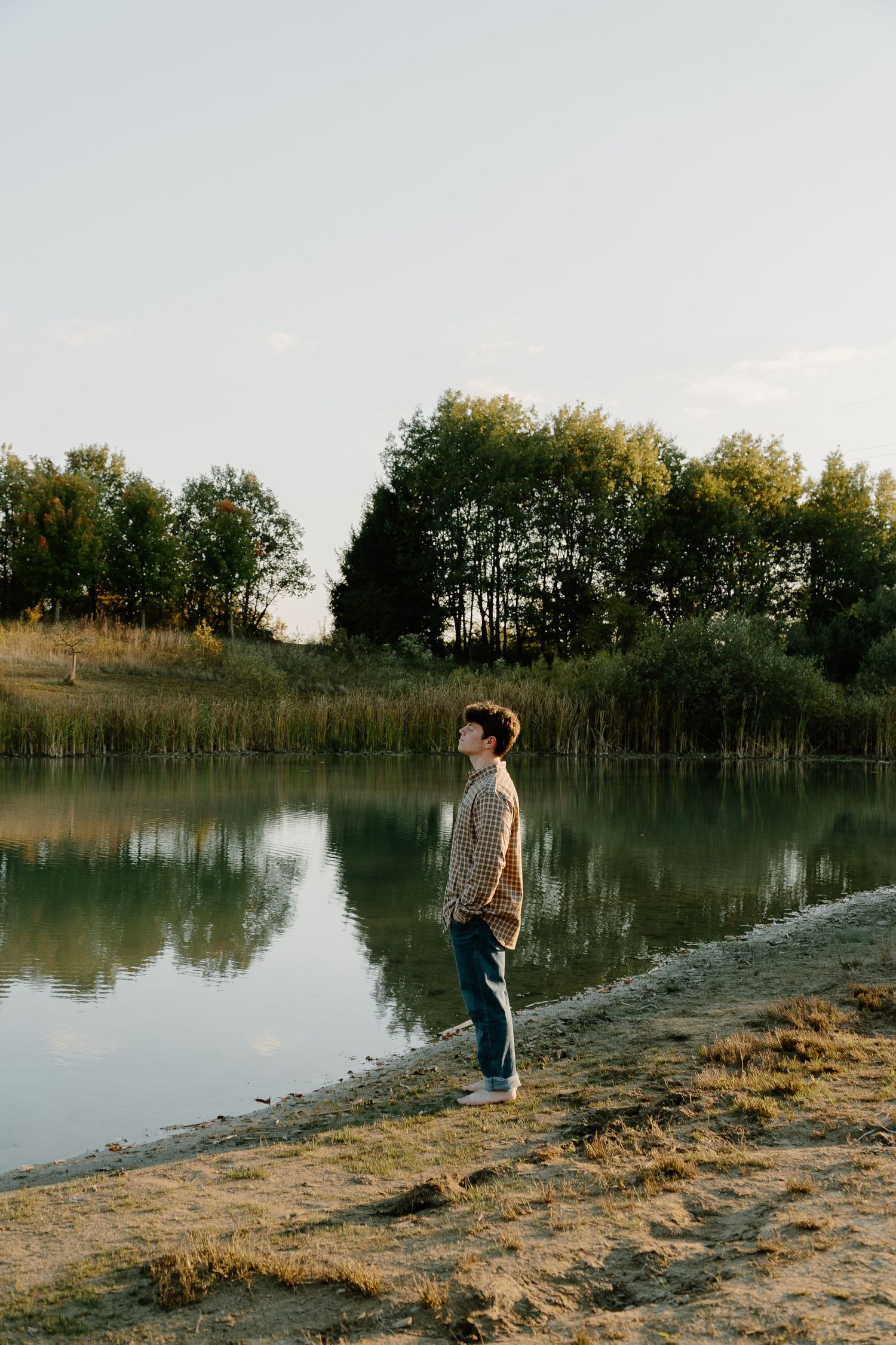 A person stands on the shore of a tranquil lake at sunset with trees reflecting in the calm water.