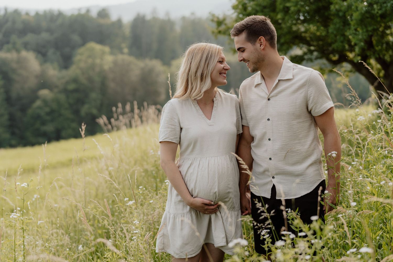 Schwangere Frau mit Partner beim Spaziergang durch eine Wiese, beide lachen sich an