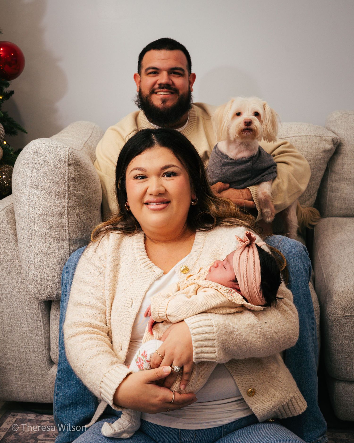 Couple relaxing on grey couch with small white dog during holiday photoshoot.